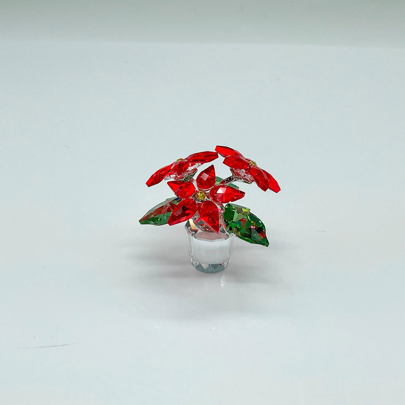 Swarovski Crystal Figurine, Poinsettia: A small Christmas flower. Red petals with a yellow center and green leaves. Resting on a clear flower pot. Swarovski marked. This item has its original box: 4.25"L x 4.25"W x 3.25"H. Artist: Elke Hube