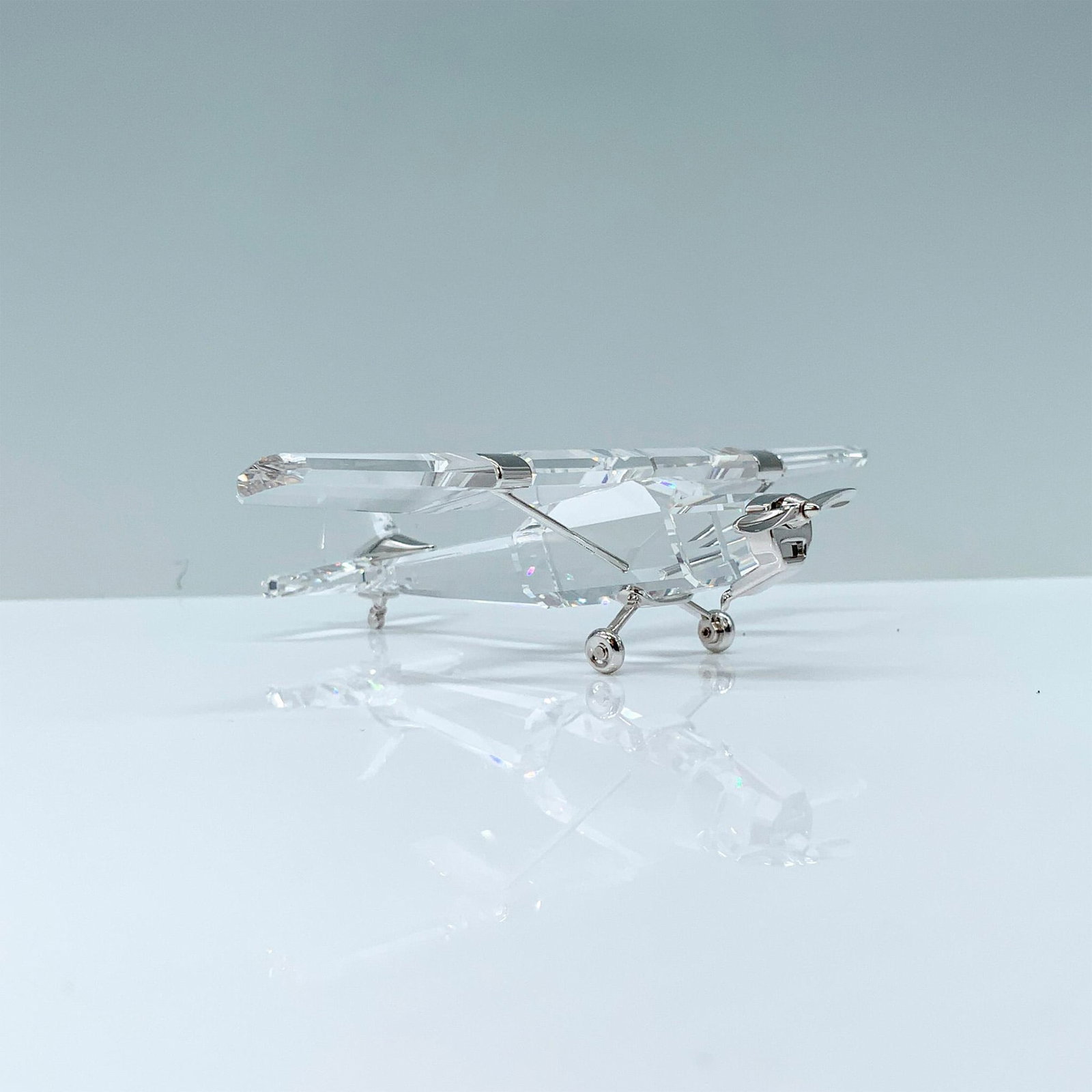 Swarovski Crystal Figurine, Rhodium Airplane: A clear crystal figure of an antique aircraft that is decorated with rhodium accents for the propeller, wheels, and struts. Swarovski backstamp. This item has its original box. Dimensions of the origi