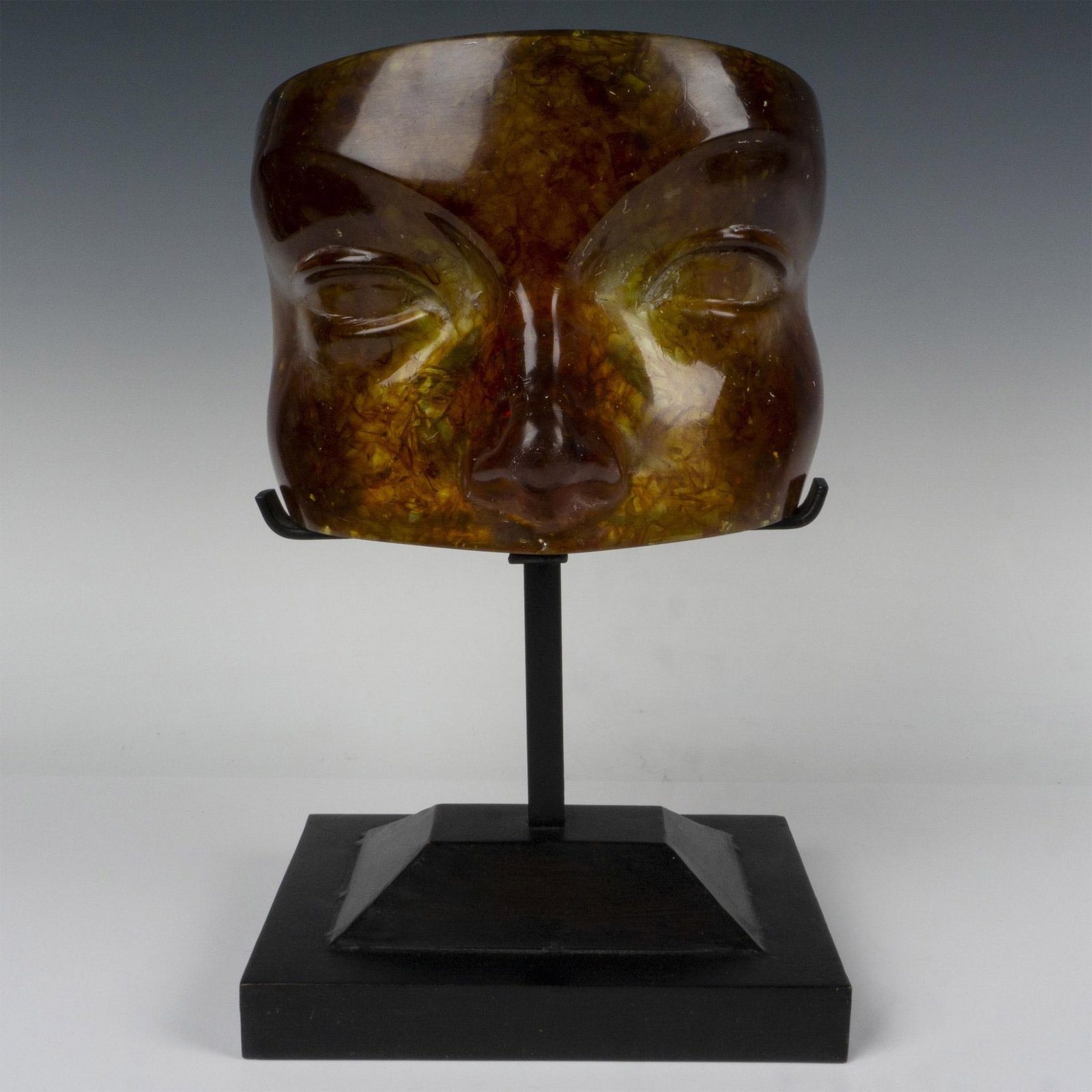 Amber Acrylic Mask Candle Holder: An amber color face sculpture formed as a person's eyes and nose. Placed on a black painted candle holder stand with a rectangular base. Dimensions: 8.25"L x 8"W x 13.75"H