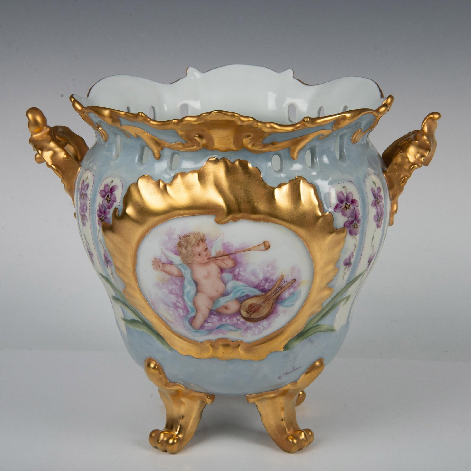 Giraud Limoges Cherub Footed Jardiniere with Purple Irises: An outstanding hand-painted antique jardiniere showcasing an angelic cherub playing a flute and guitar. The piece features a reticulated collar with a gold rim, gold double handles, and gold pedestal 