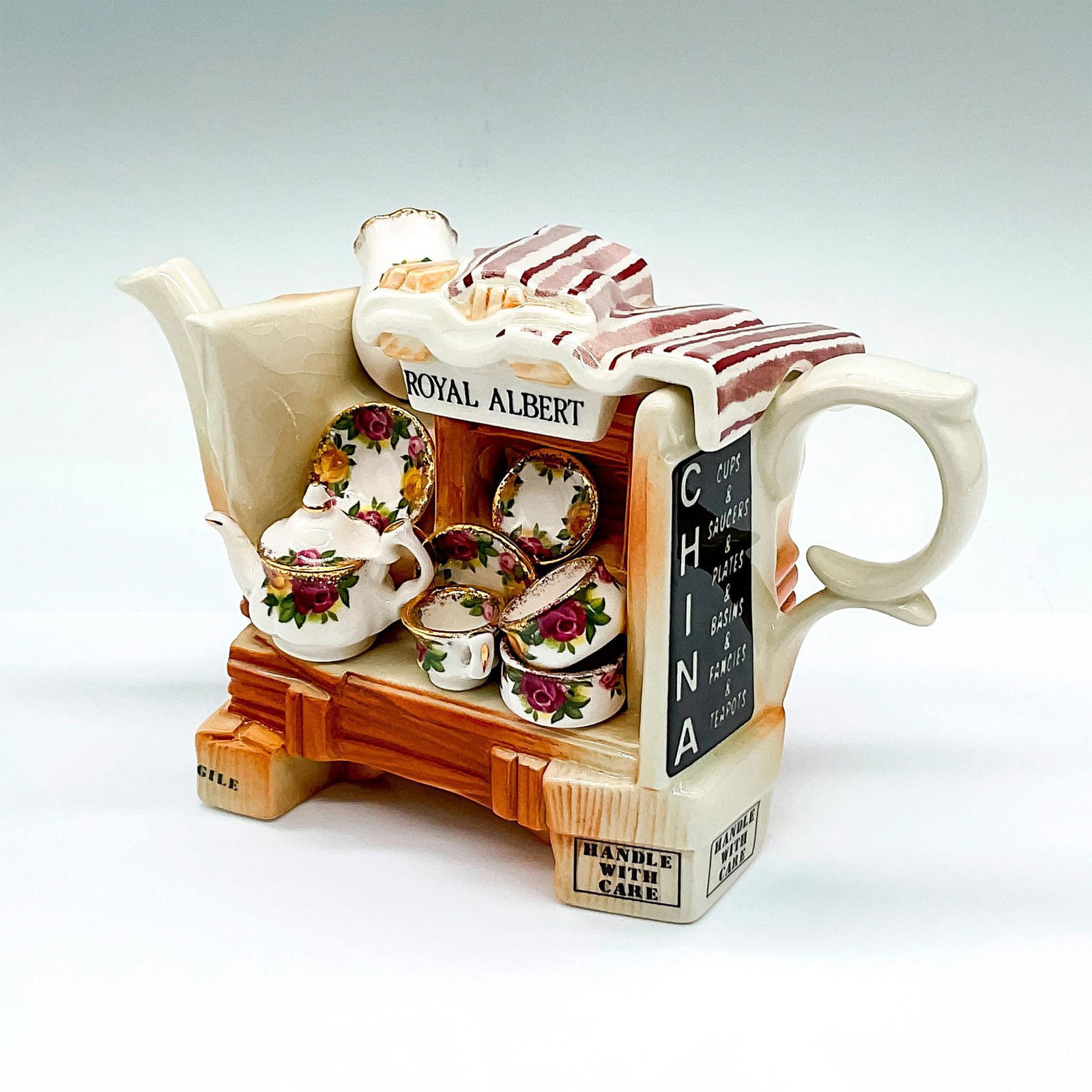 Royal Albert Earthenware Teapot, Old Country Roses: A white and brown decorative teapot molded to imitate a shop cart with a mini tea set, basin, plate, and vase in Old Country Roses pattern with gilt trims. The lid is molded as the cloth top of the ca