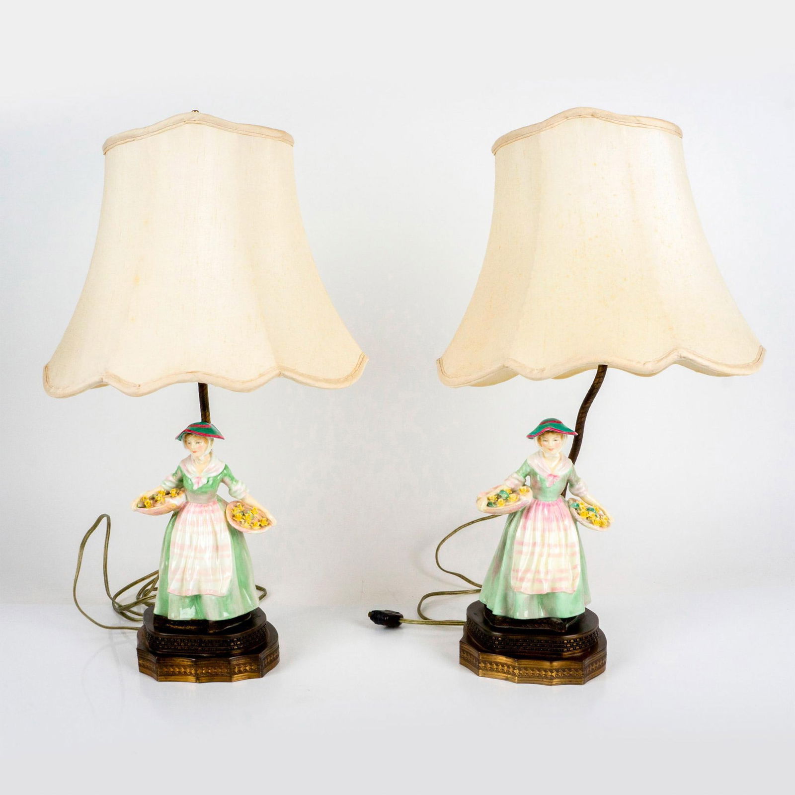 Pair of Daffy Down Dilly HN1712 Table Lamps by Royal Doulton: HN1712 figure of a daffodil seller in green; attached to a brass base. Includes cream color lampshades and lightbulbs. Artist: Leslie Harradine Dimensions: 13"L x 9"W x 22"H Manufacturer: Royal Doulto