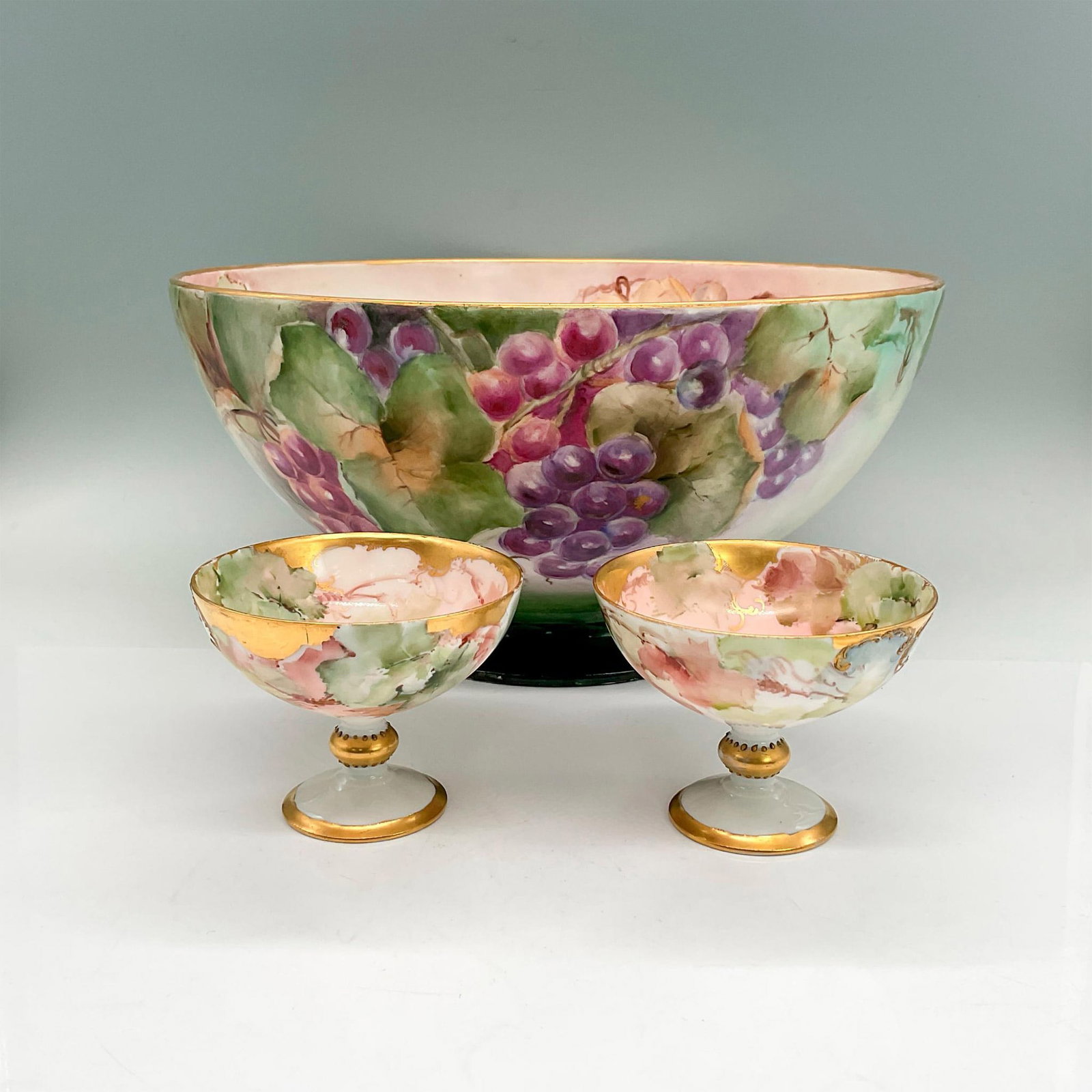 3pc W.G. & Co. Limoges Porcelain Bowl Grapes + 2 Cups: Large footed bowl with hand-painted purple and green grapes, green leaves, gold rim; 6"H x 12" dia. Signed M.E. Clare. W.G. & Co. backstamp. Two pedestal cups 3"H x 3.75" dia; J.P.L. backstamp. Issued