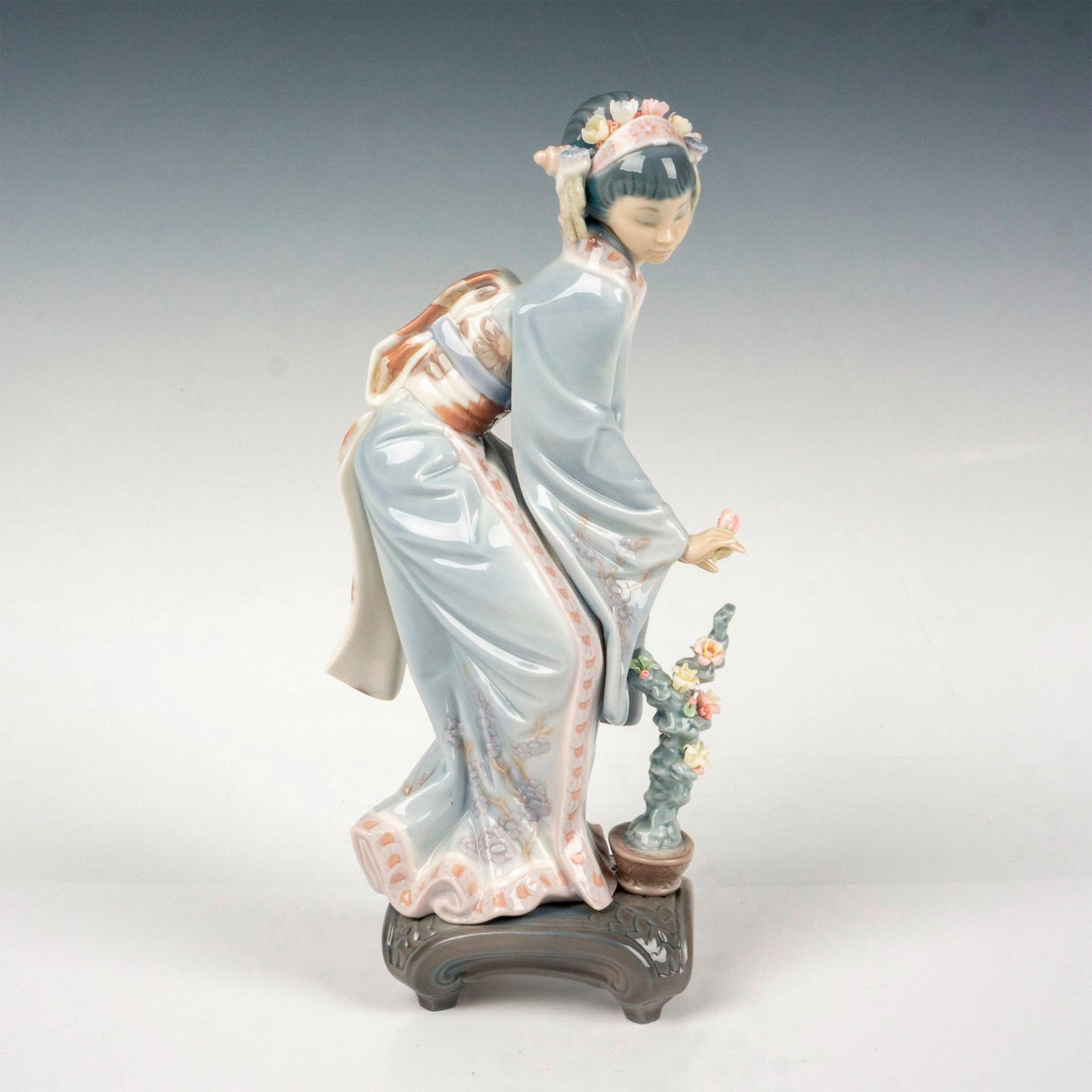 Lladro Porcelain Figurine, Mayumi 1001449: A glossy figure of a Japanese woman in a floral kimono over a potted plant. Lladro backstamp. This item has its original box: 12.5"L x 7.25"W x 6"H. Artist: Salvador Debon Issued: c. 1983-1998 Dimensi
