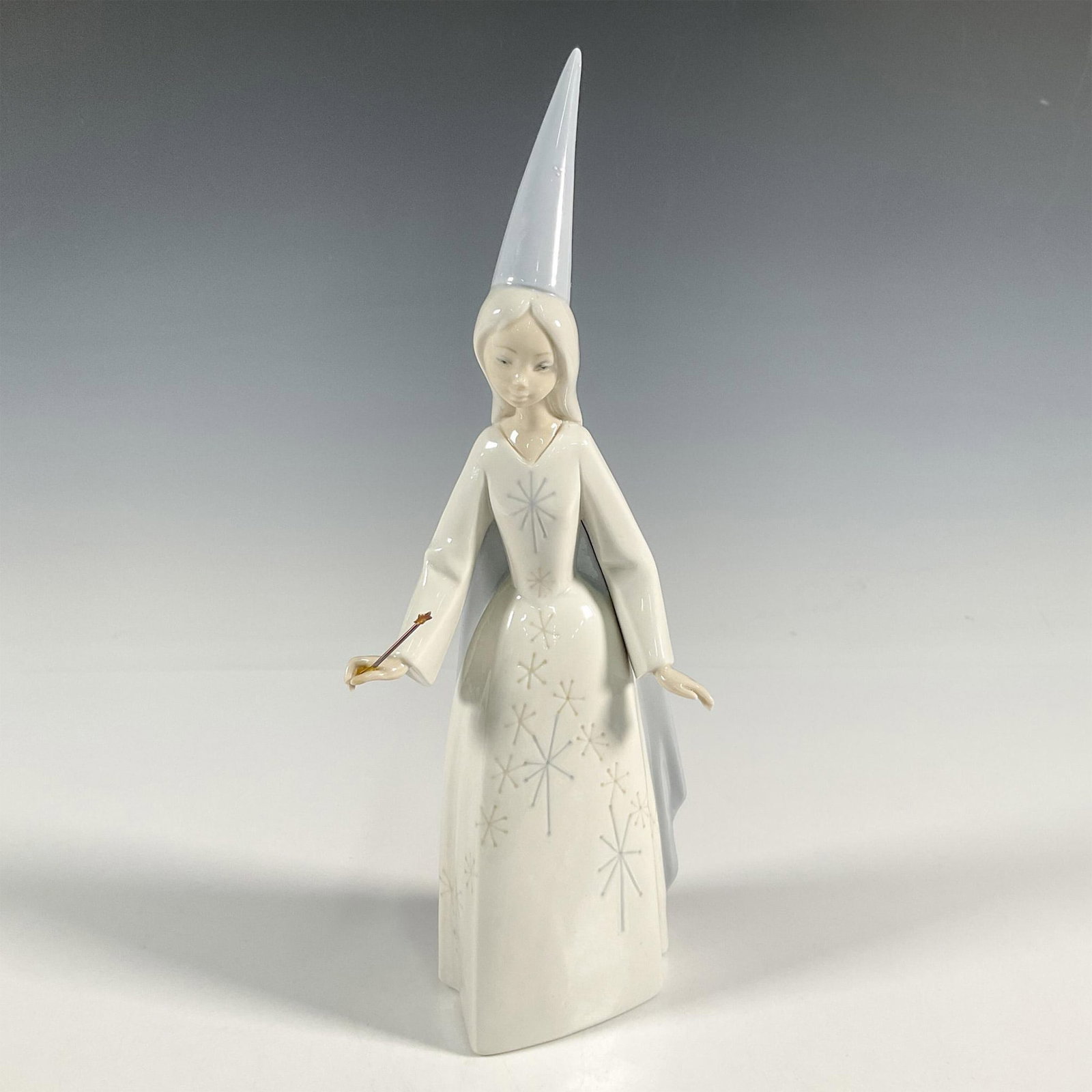 Fairy 1004595 - Lladro Porcelain Figurine: A glossy figure of a young fairy in a blue pointed hat and long cape. She is holding a metal star wand. Lladro backstamp. This item has its original box: 12.5"L x 7.5"W x 6"H. Artist: Vincente Martine