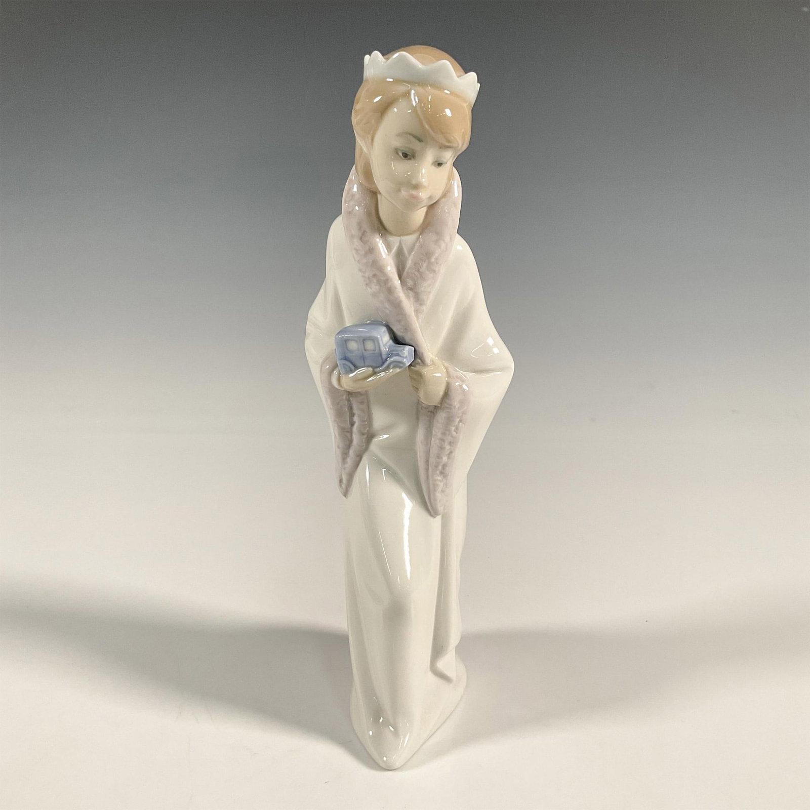 King Gaspar 1004674 - Lladro Porcelain Figurine: A glossy figure of a girl dress as a king holding a blue toy car. Lladro backstamp. Artist: Juan Huerta Issued: 1969-2001 Dimensions: 4.25"L x 3"W x 8"H Manufacturer: Lladro Country of Origin: Sp