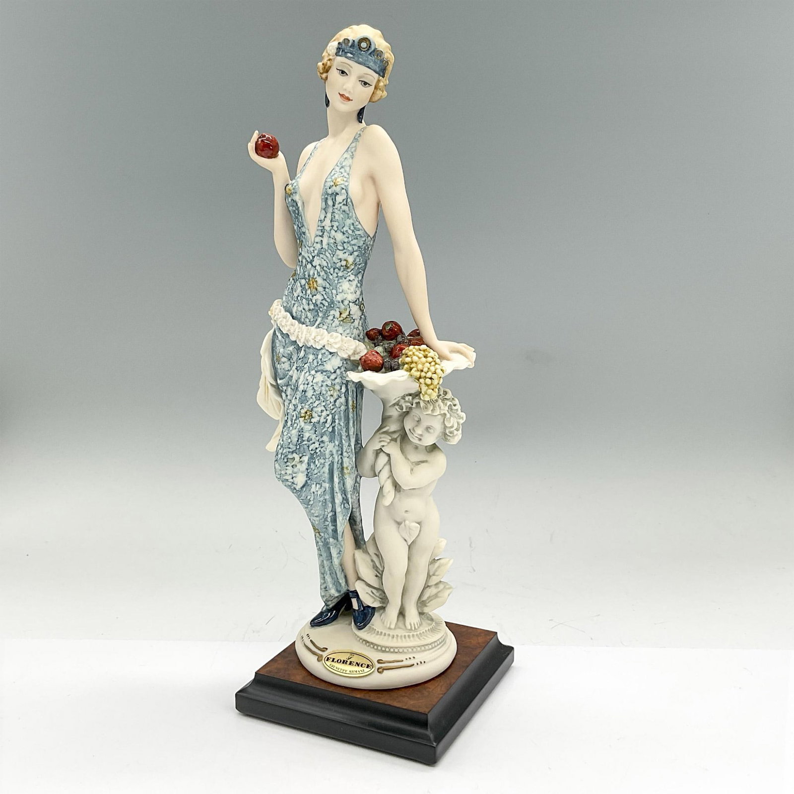 Florence Sculture d'Arte Giuseppe Armani Figurine, Indigo: Cream colored matte figure of a stylish Art Deco era lady in a blue patterned dress, leaning on a cornucopia topped statue while holding an apple. Florence Sculture d'Arte affixed label. Artist: Giuse