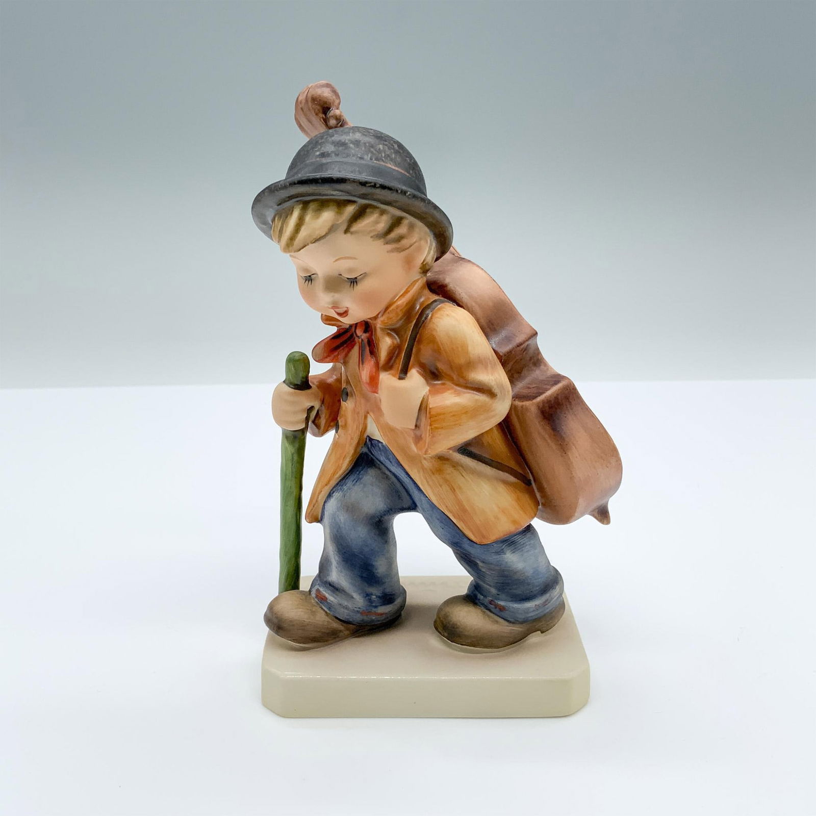 Little Cellist #89/I - Goebel Hummel Figurine: Hand-painted figurine depicting a boy with a walking stick and large cello on his back. TMK-6 Goebel backstamp on the bottom. Issued: c. 1991 Dimensions: 3"L x 2"W x 5.5"H Manufacturer: Goebel Hummel