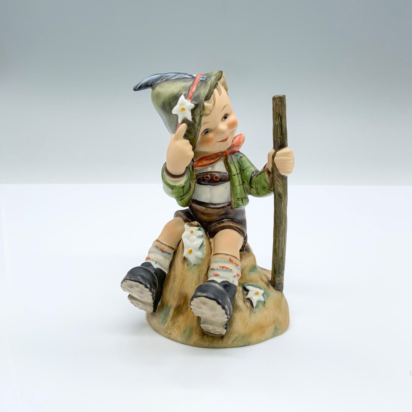 Mountaineer 315 - Goebel Hummel Figurine: Hand-painted figurine depicting a boy sitting with a walking stick pointing at a flower on his hat. Goebel backstamp on the bottom. Dimensions: 3"L x 2.75"W x 5.25"H Manufacturer: Goebel Hummel Countr