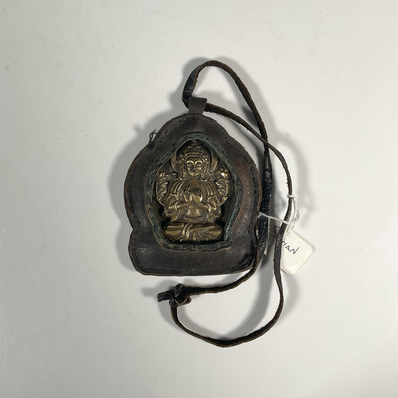 Tibetan Travel Bronze Buddha Deity Encased in Leather: Modeled metal plaque of Buddha in prayerful seated pose. Pendant: 3.75"W x 4.5"H. Lanyard only: 14.75"L. Issued: 20th century