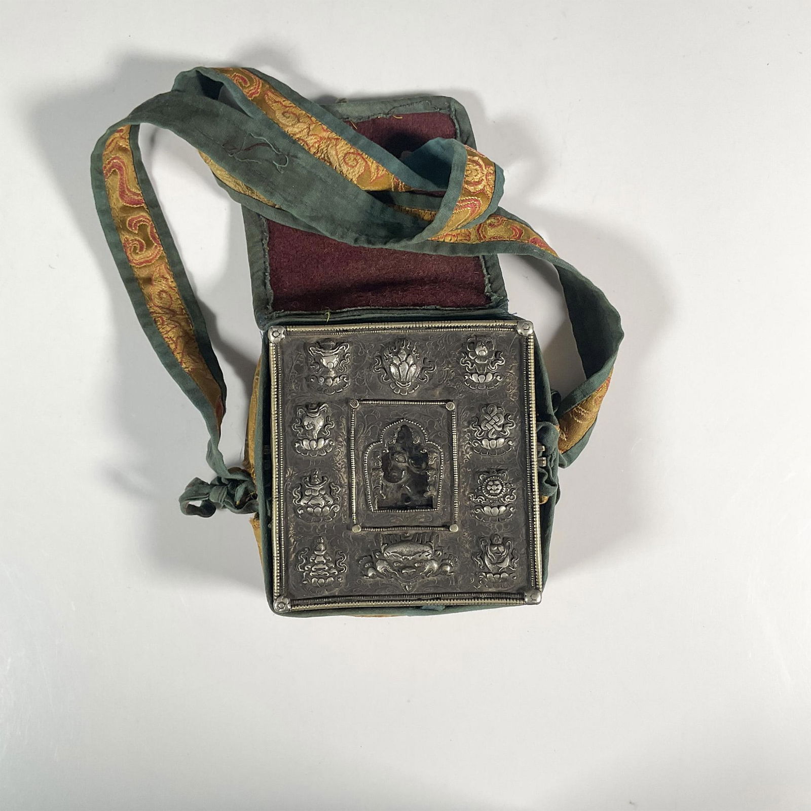 Tibetan Traveling Shrine Box: A metal box that contains a figure of a protector deity inside, decorated with repousse flowers and a dragon face. The box is carried in a decorative fabric case with strap. Box in pouch: 5.75"L x 2"W