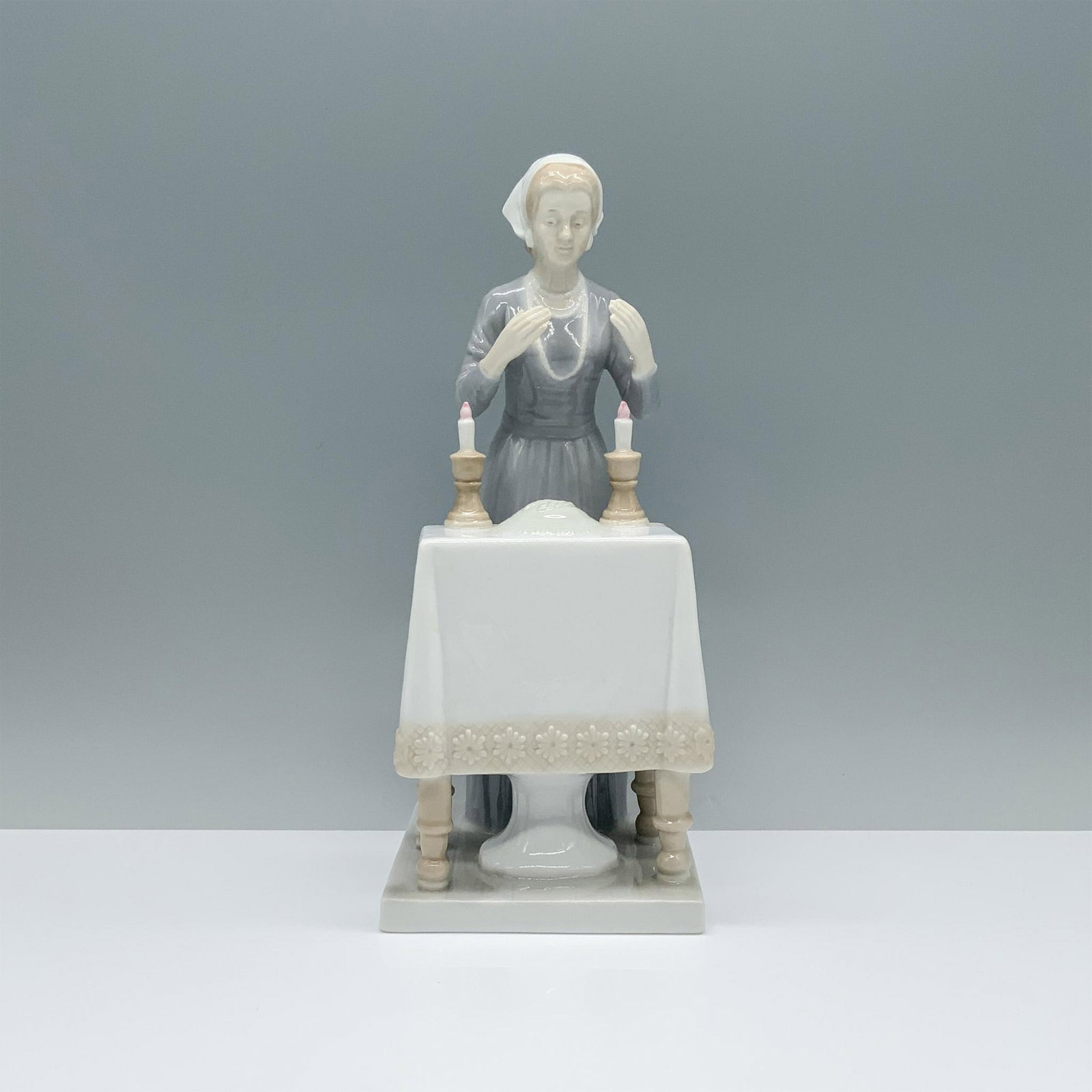 Moshe Yakov Judaica Candle Lighting Figurine: Capturing timeless elegance, a woman in a gray dress stands gracefully at a prepared table adorned with two lit candles. She wears a pearl necklace and a white headscarf, her hands raised in a moment 