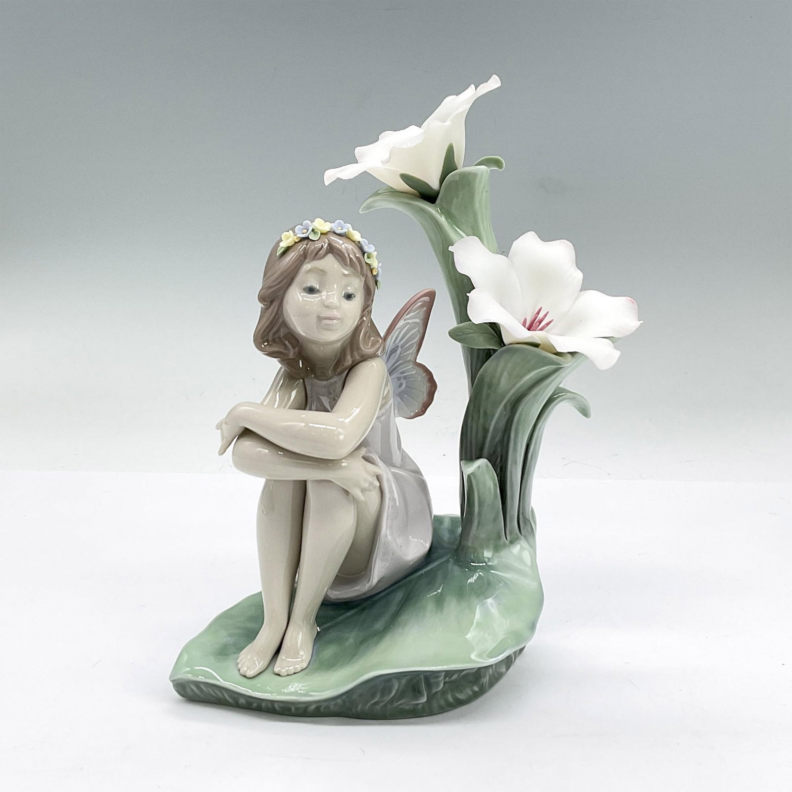 Lladro Porcelain Figurine, Lakeside Daydream 1006644: Glazed porcelain figure of a young fairy sitting on a leaf with flowers next to her. Lladro backstamp. This item has its original box: 10"L x 9"W x 6.25"H. Issued: 2000-2007 Dimensions: 5"L x 3.75"W x