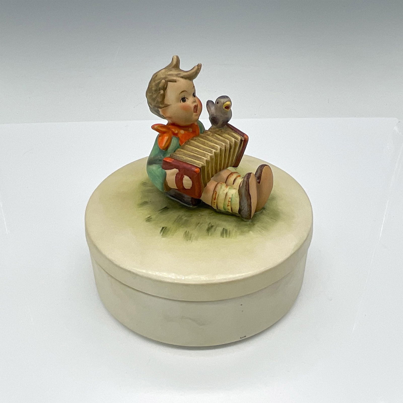 Goebel Hummel Figural Powder Box, Let's Sing HUM III/110: Hand-painted lidded powder box with a figure of a boy sitting on the top lid, depicted with his accordion and bird friend. Goebel TMK3. Issued: 1960-1972 Dimensions: 5.25"H x 5.5" dia. Manufacturer: G