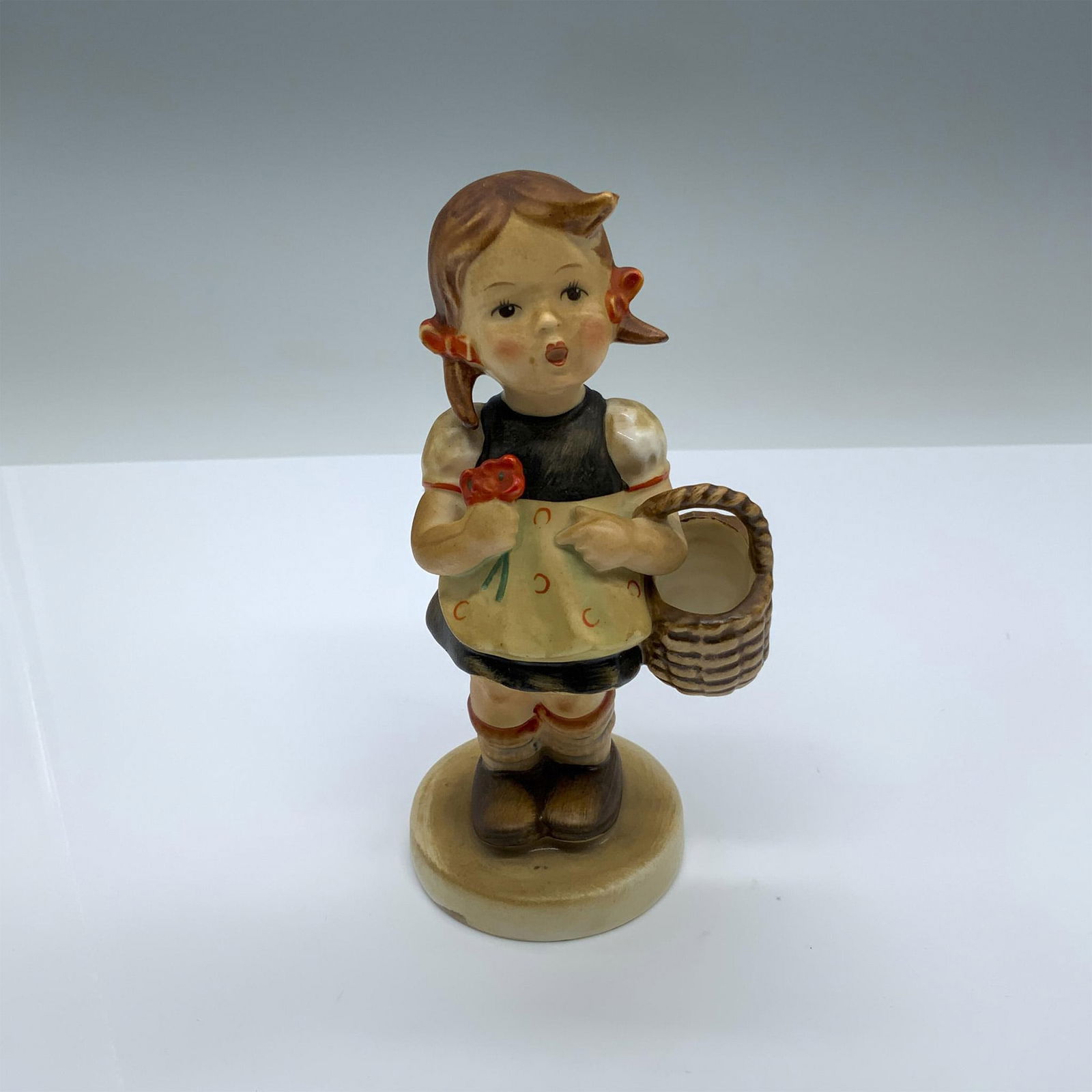 Goebel Hummel Porcelain Figurine, Girl With Basket 98: Hand-painted figurine depicting a young girl holding a basket and flowers. Goebel backstamp on the bottom. Dimensions: 3"L x 2.5"W x 5.75"H Manufacturer: Goebel Hummel Country of Origin: Germany