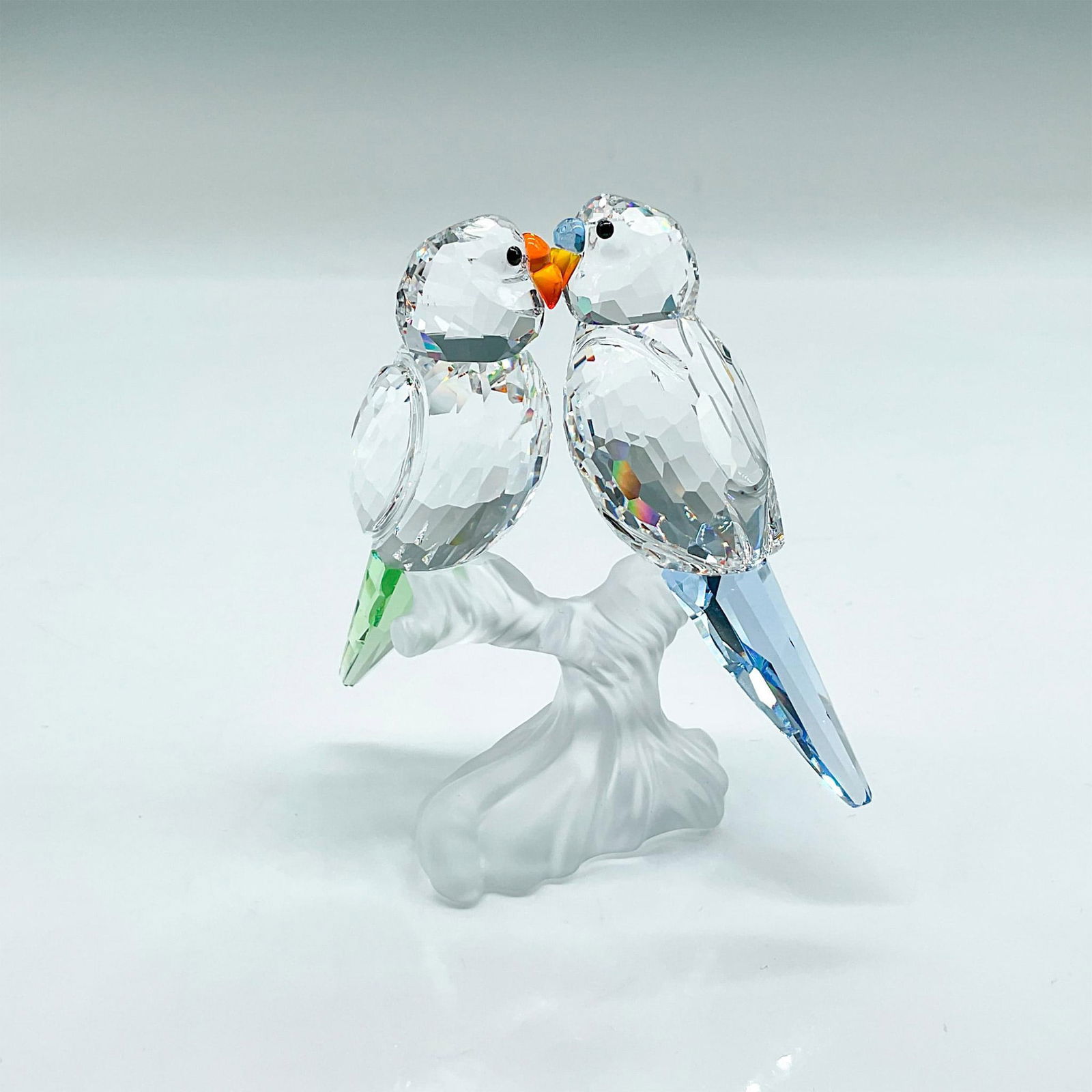 Swarovski Crystal Figurine, Pair of Budgies: Clear faceted crystal figural of two budgies perched on a frosted base. One has light blue tail feathers, the other light green. Swarovski etched backstamp. Includes original box 6"L x 6"W x 4"H. Issu