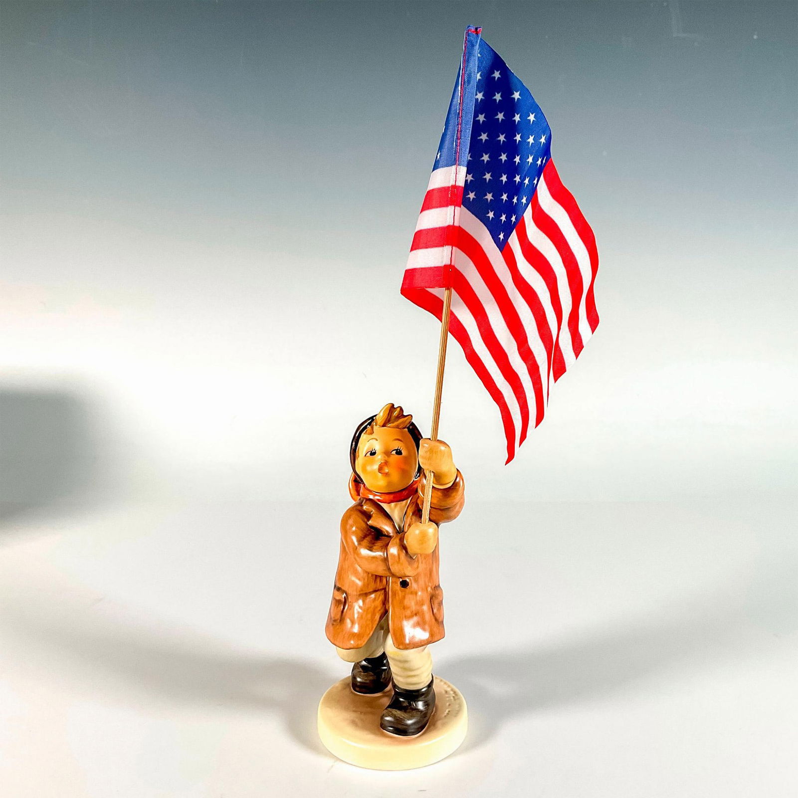Goebel Hummel Figurine, Call To Glory: Hand painted in matte finish. Boy proudly holds a flag in hand. Includes an American flag, a German flag, and the flag of Europe. Goebel backstamp. This item has its original box. Box measures: 5"L x