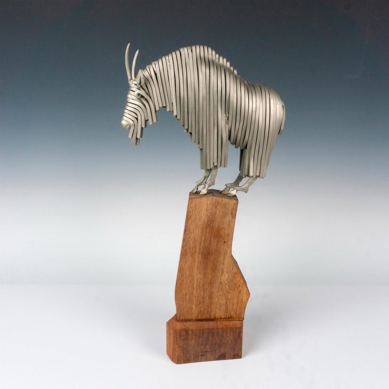Fay Taylor Billy Goat Metal Sculpture on Wood Base: An engaging metal sculpture crafted from multiple fused metal lengths, forming a captivating goat atop a tall wood base. The composition gives the impression of the goat perched on a hilltop, creating