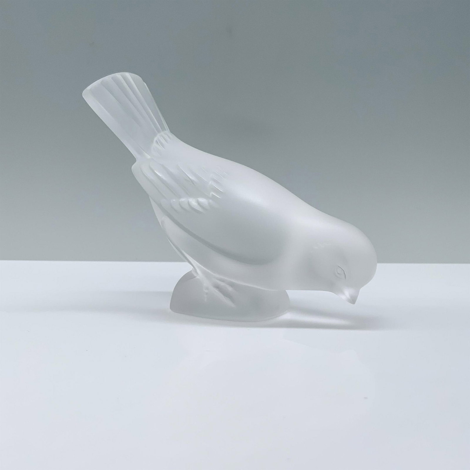Lalique Crystal Figurine, Moineau Hardi: A frosted crystal figurine portrays a sparrow with its head inclined towards the dirt, as if inquisitively peering downward, with a raised backside. Signed Lalique France. Dimensions: 5"L x 2.25"W x 3
