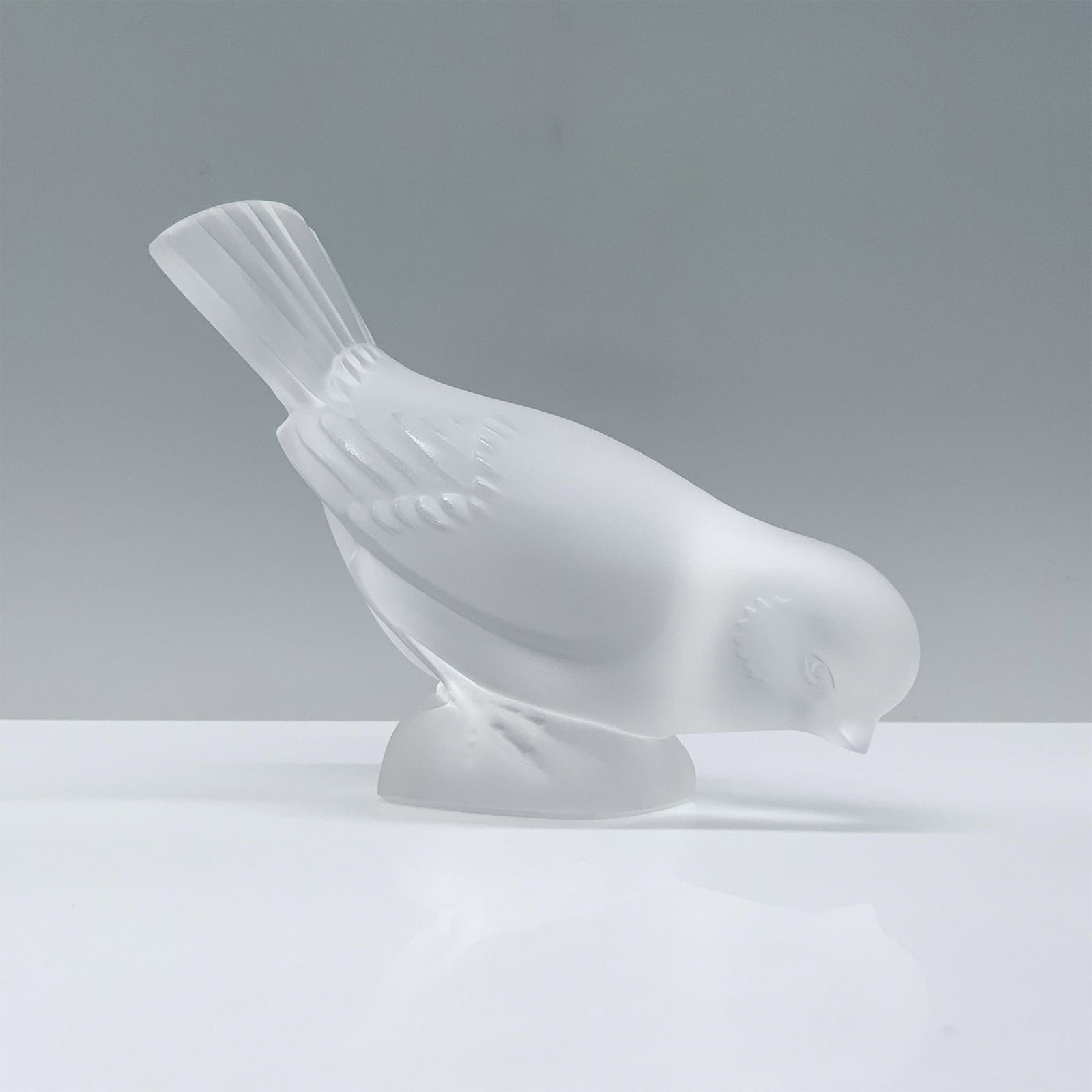 Lalique Crystal Figurine, Moineau Hardi: A frosted crystal figurine portrays a sparrow with its head inclined towards the dirt, as if inquisitively peering downward, with a raised backside. Signed Lalique France. Dimensions: 5"L x 2.25"W x 3