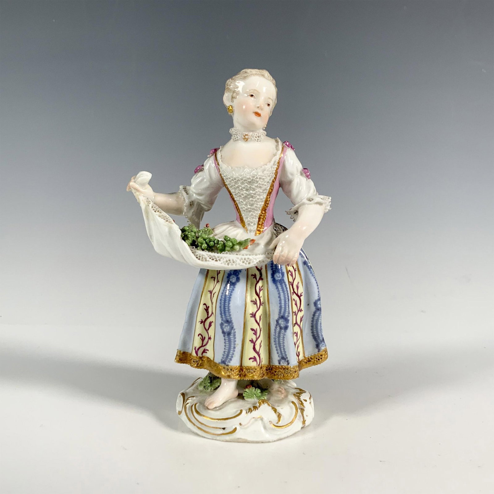 Meissen Style Porcelain Woman with Grapes Figurine: German porcelain figure of woman selling grapes. She wears an 18th century costume, with grapes laid out on cloth, standing on gilded base. Marked to base. Dimensions: 3.5"L x 2.25"W x 5.5"H
