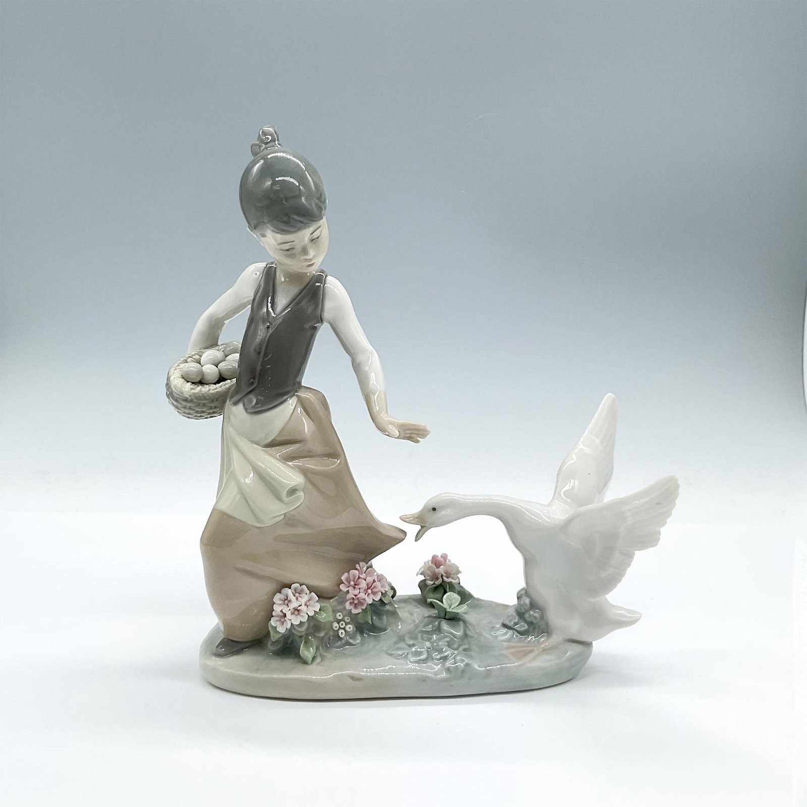 Lladro Porcelain Figurine, Aggressive Goose 1001288: Lovely young girl with her basket of eggs shooing a goose. Pretty flowers adorn the ground. Glossy finish. Lladro backstamp. This item has its original box. 12.25"L x 10"W x 5.75"H. Artist: Juan Huert