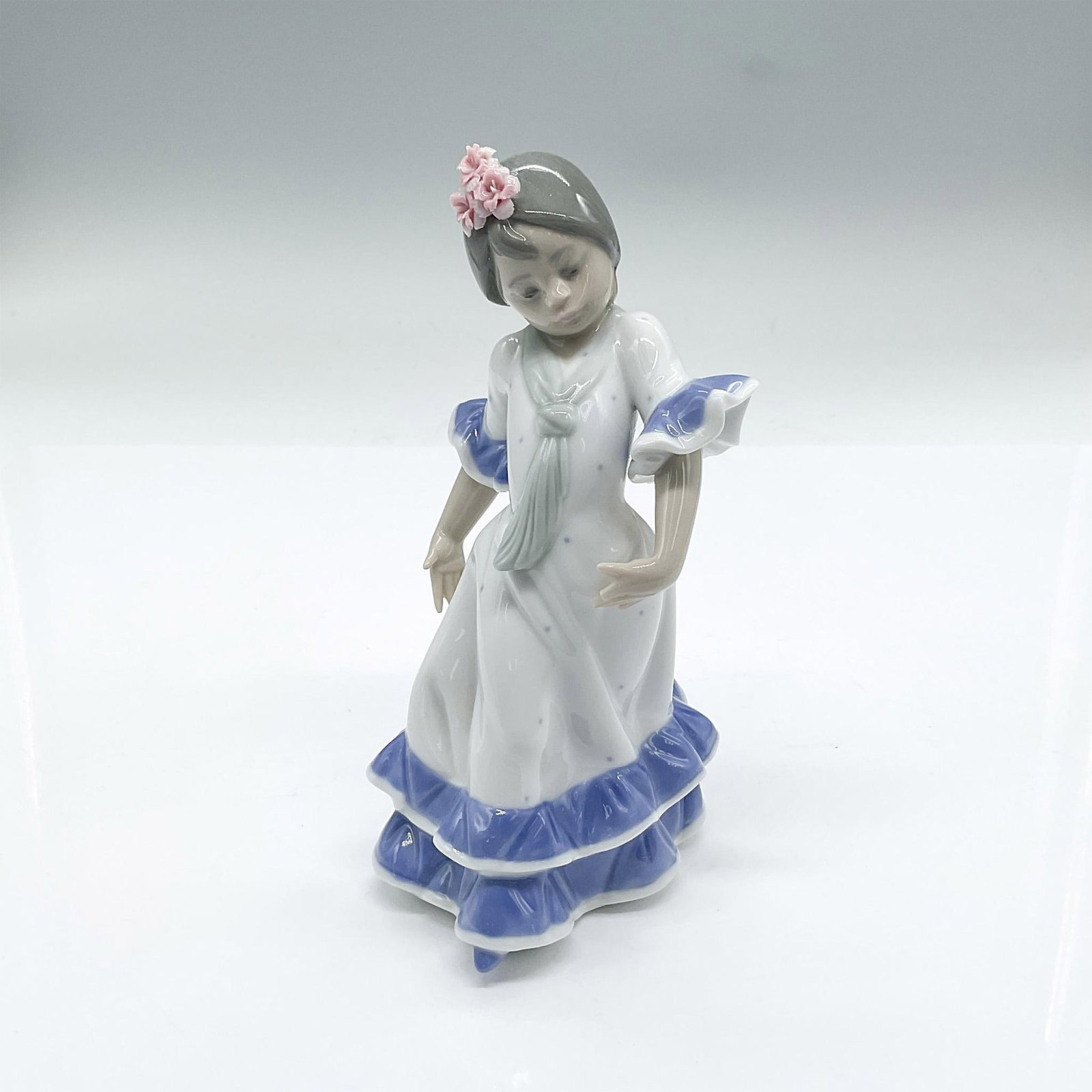 Lladro Porcelain Figurine, Juanita: In a ruffled white dress with blue trim. Gloss finish. Lladro backstamp. Issued: 20th century Dimensions: 3.5"L x 4"W x 6.75"H Manufacturer: Lladro Country of Origin: Spain