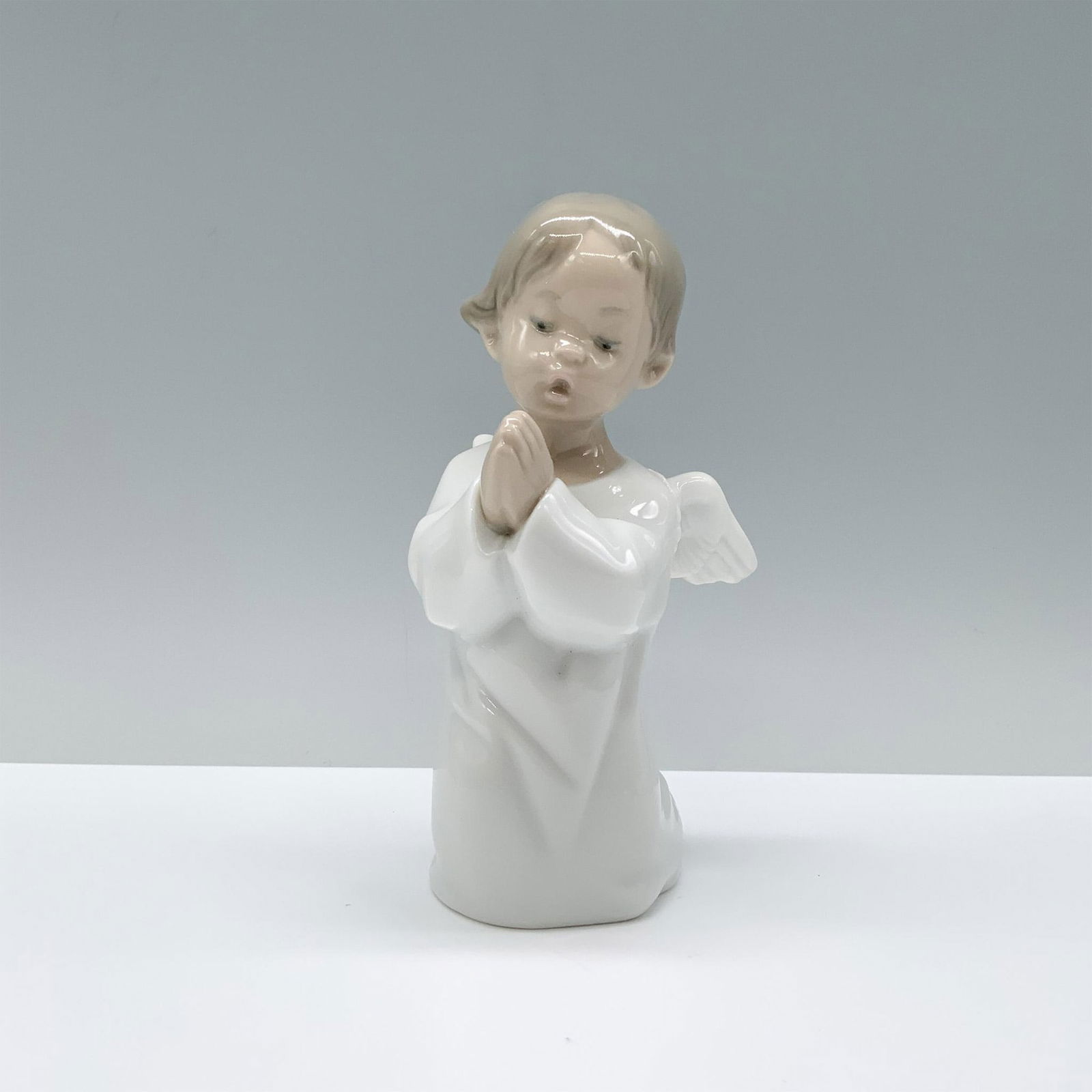 Lladro Porcelain Figurine, Praying Angel 1004538: A figurine of a young angel child in serene prayer, adorned in a pristine white robe with graceful, open wings. Lladro backstamp. Artist: Fulgencio Garcia Issued: 1969 Dimensions: 3"L x 3"W x 5.25"H M