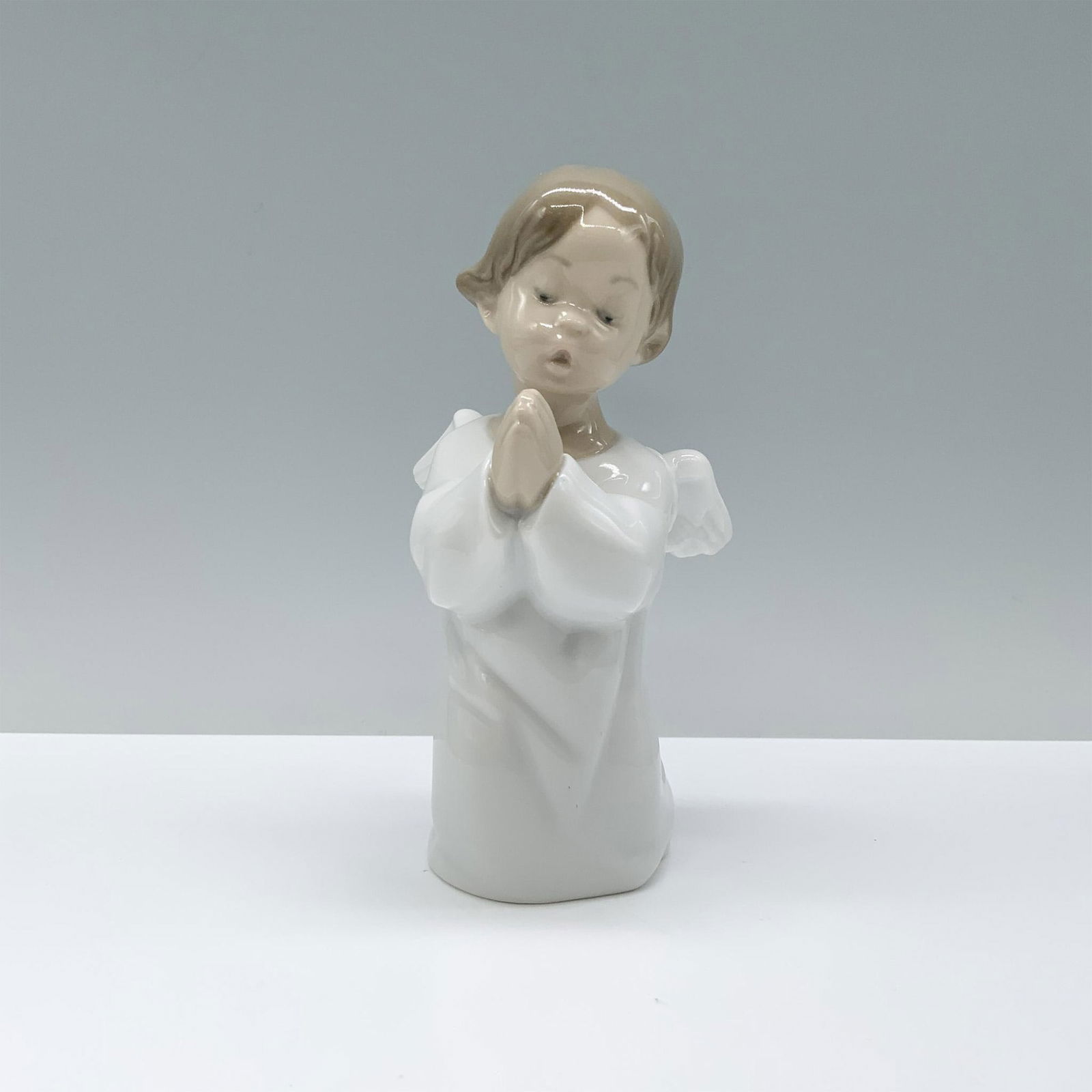 Lladro Porcelain Figurine, Praying Angel 1004538: A figurine of a young angel child in serene prayer, adorned in a pristine white robe with graceful, open wings. Lladro backstamp. Artist: Fulgencio Garcia Issued: 1969 Dimensions: 3"L x 3"W x 5.25"H M