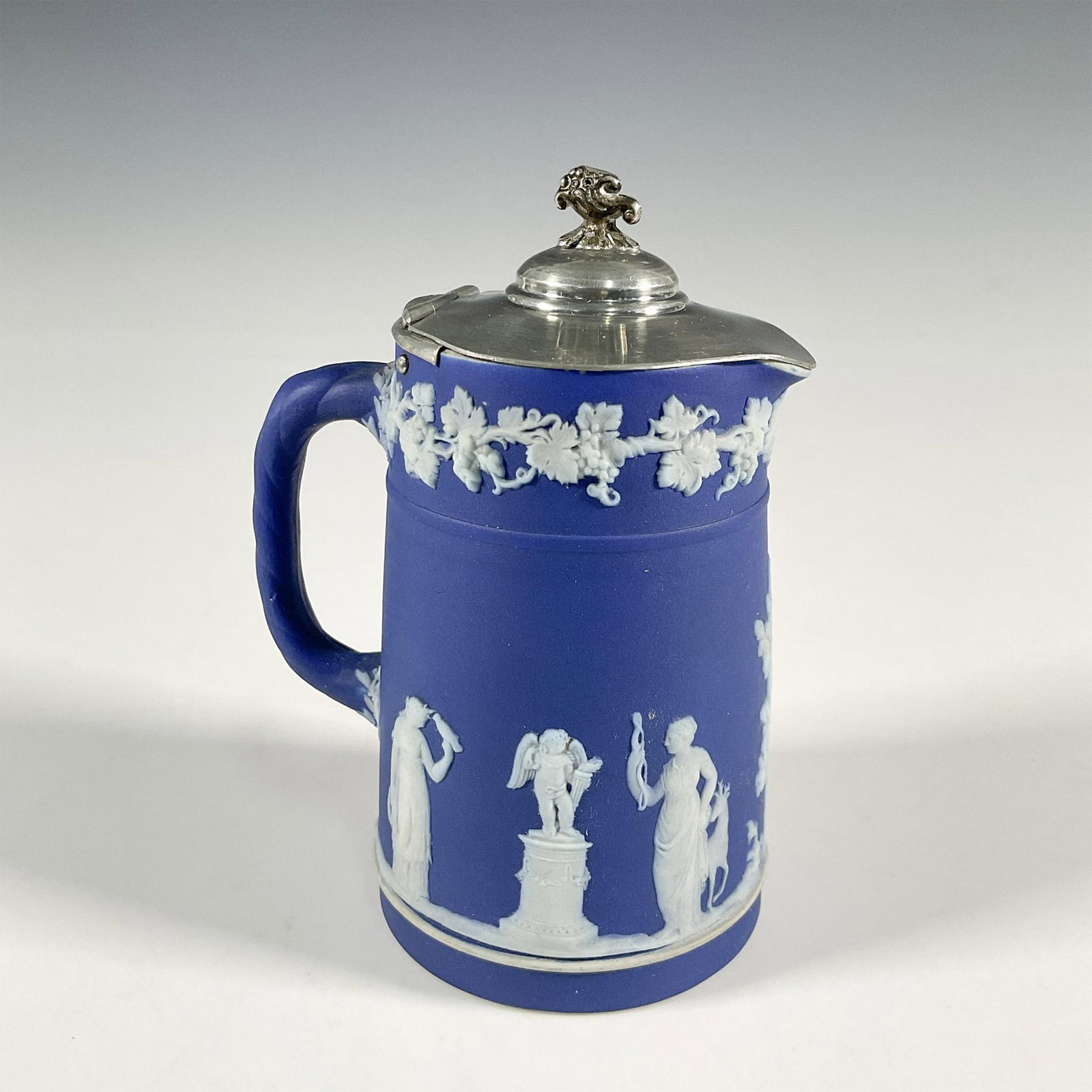 Wedgwood Blue Jasperware Jug with Silverplate Lid: Blue jasperware jug with silver plate lid, floral finial on lid. The jug is decorated in relief with white neoclassical designs. Wedgwood impressed backstamp. Dimensions: 3.5"L x 2.75"W x 5.25"H Manuf