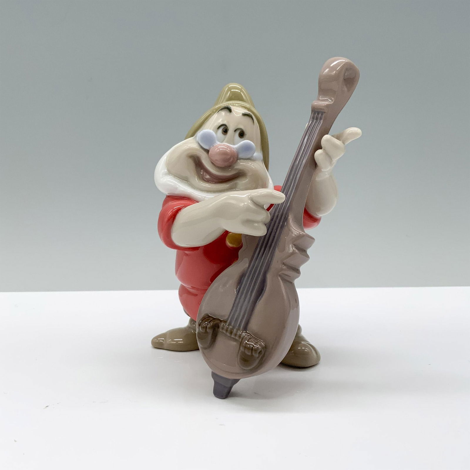 Nao by Lladro Porcelain Disney Figurine, Doc: Bring joy to your collection with Doc from Snow White, dressed in his traditional outfit, smiling brightly as he holds a guitar in his hands. Nao by Lladro backstamp. Artist: Eva Maria Cuerva Issued: