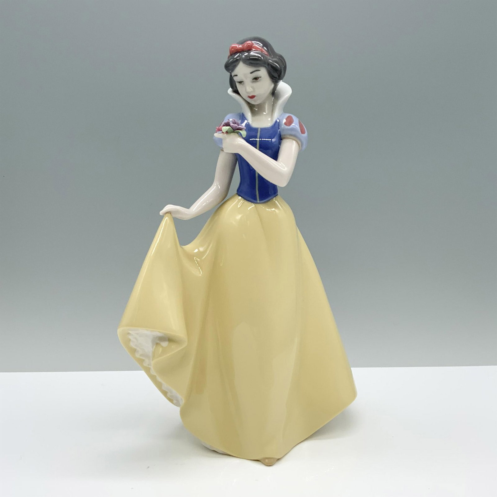 Nao by Lladro Porcelain Disney Figurine, Snow White: Capture the timeless grace of Snow White with this exquisite porcelain figurine, adorned in her iconic outfit, delicately lifting her skirt with one hand while cradling a bouquet of flowers with the o