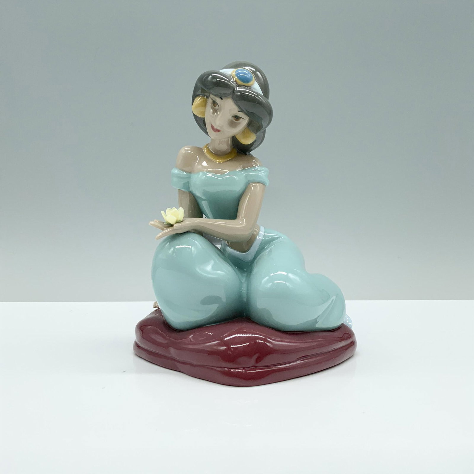 Nao by Lladro Porcelain Disney Figurine, Jasmine: A delicate porcelain figurine of Disneys Jasmine, dressed in her classic outfit, elegantly seated on crossed legs atop a pillow, captivated by a beautiful blooming flower. Nao by Lladro backstamp. Art