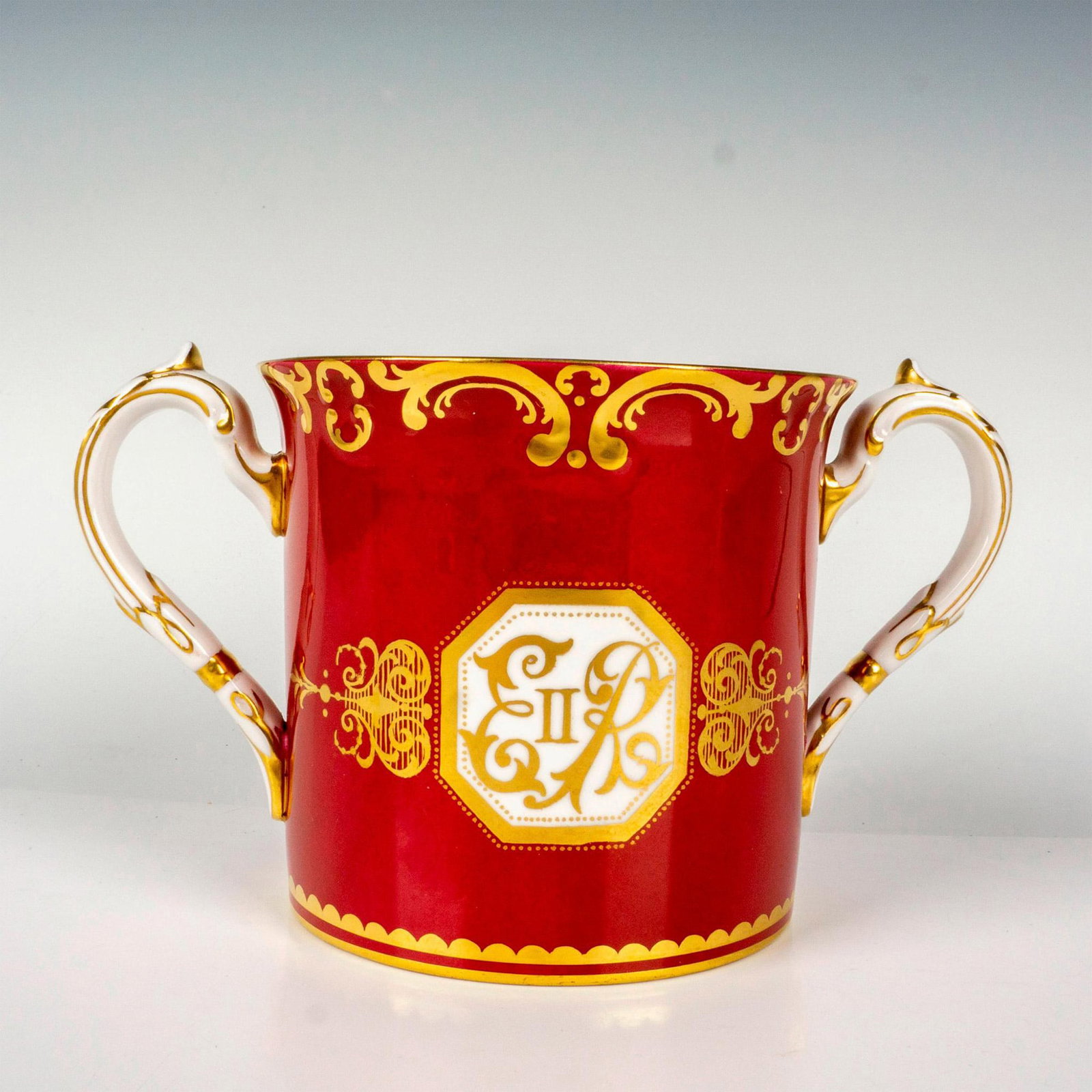 Royal Albert Commemorative Cup, Queen Elizabeth II: A commemorative bone china two-handled mug with rich red ground decorated with transfer-printed image of the Queen. Heavy gilt decoration. Royal Albert backstamp. Dimensions: 7.5"W x 4.5'H Manufacture