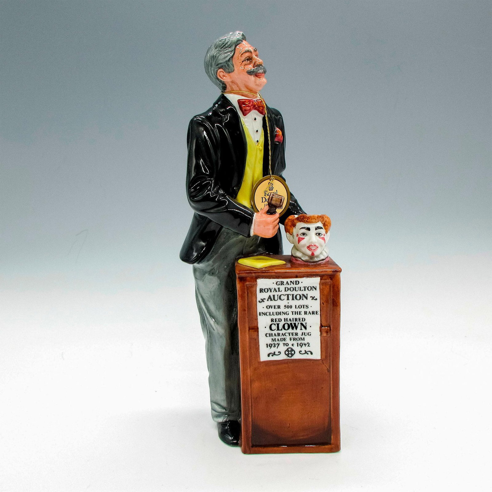 The Auctioneer - HN2988 - Royal Doulton Figurine: Glossy figural holding a red clown mug up for auction. Part of the International Collectors Club. Royal Doulton backstamp. Issued: 20th century Dimensions: 3.5"L x 3"W x 9"H Manufacturer: Royal Doulto