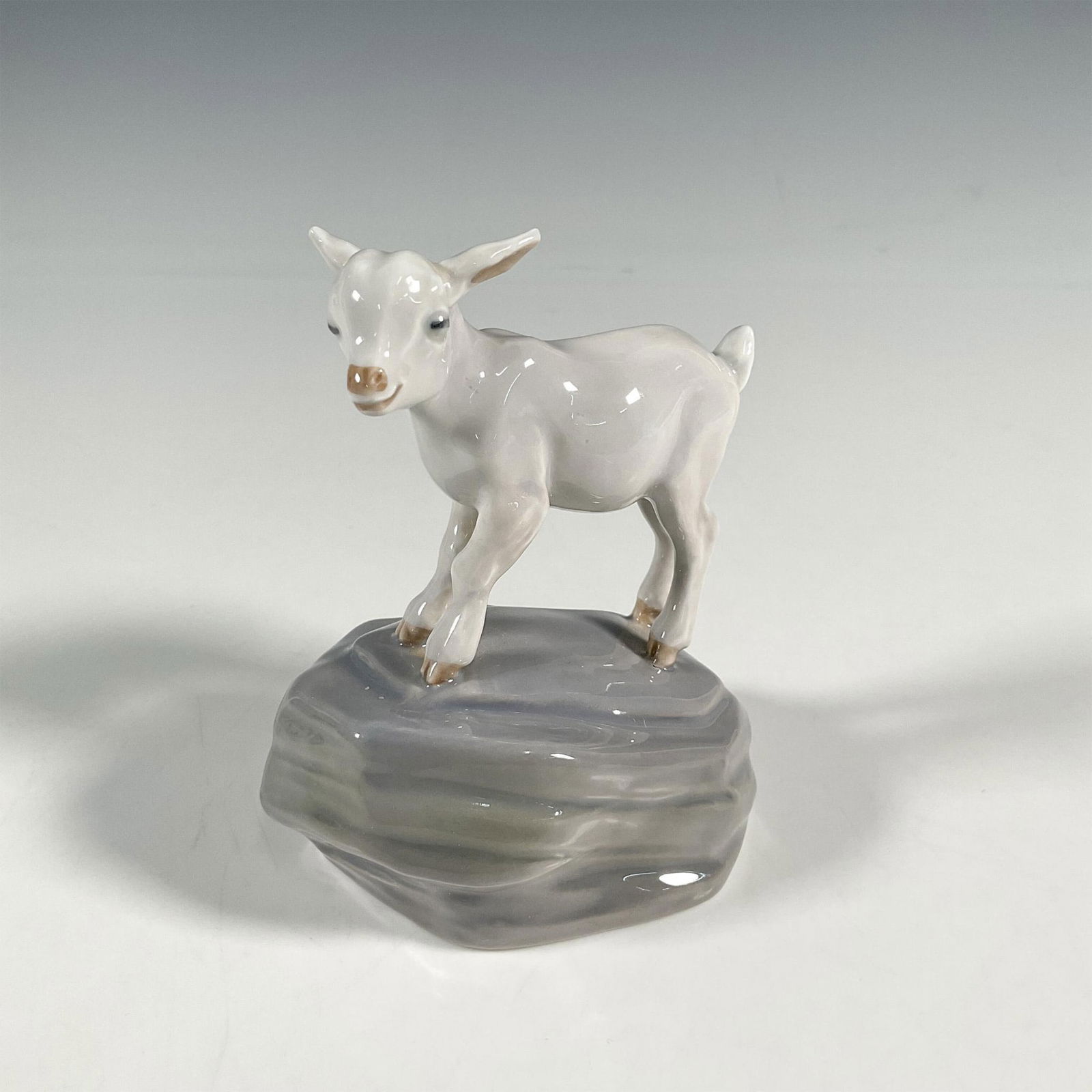 Royal Copenhagen Figurine, Goat Kid On Rock: Glazed finish figure of goat kid standing on a rock. Royal Copenhagen backstamp. Artist: Jeanne Grut Dimensions: 2.5"L x 3.5"W x 4"H Manufacturer: Royal Copenhagen Country of Origin: Denmark