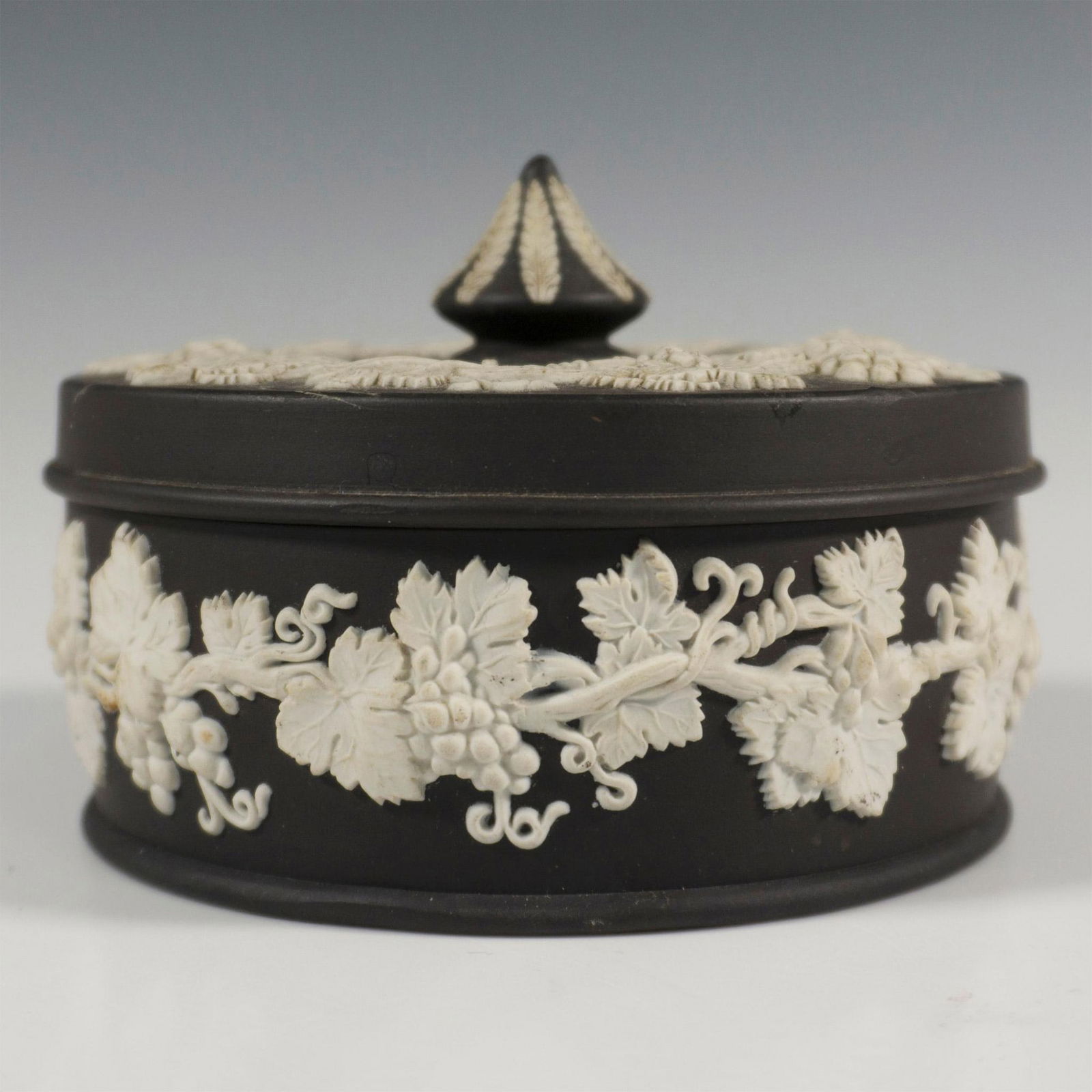 Wedgwood Black Jasperware Covered Candy Bowl: Black and white jasperware candy bowl with lid of round form, decorated in a relief depicting grapevine motif encircling the lid and body. Wedgwood impressed backstamp. Dimensions: 4.75"W x 3.75"H Man