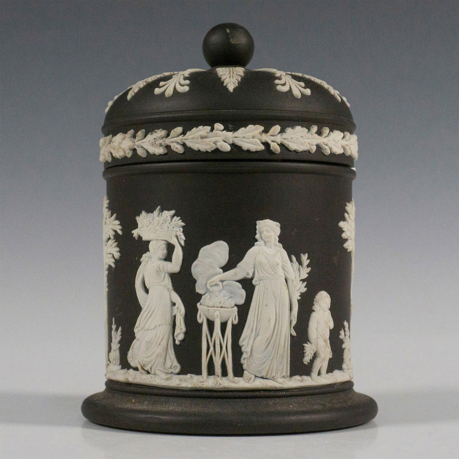 Wedgwood Black Jasperware Lidded Tobacco Jar: Black and white canister with lid, decorated in a relief with classical figures offering sacrifices with acanthus leaf and fan palms. Wedgwood impressed backstamp. Dimensions: 3.25"W x 5"H Manufacture