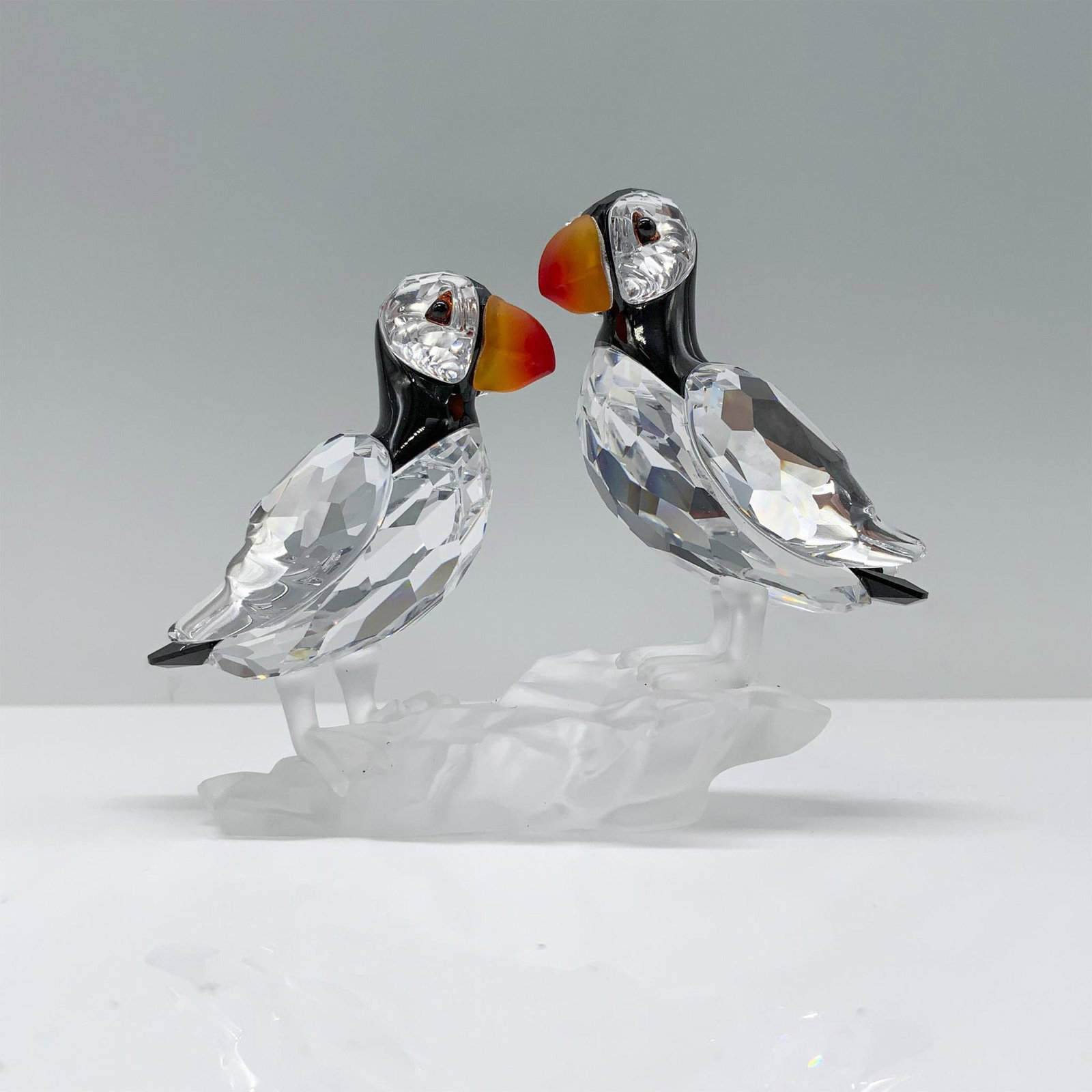 Swarovski Crystal Figurine, Puffins 261643: A charming pair of clear crystal puffins, with sleek black crystal heads and backs and vibrant red and yellow gradient beaks, while perched atop frosted crystal rocks. This item has its original box. 