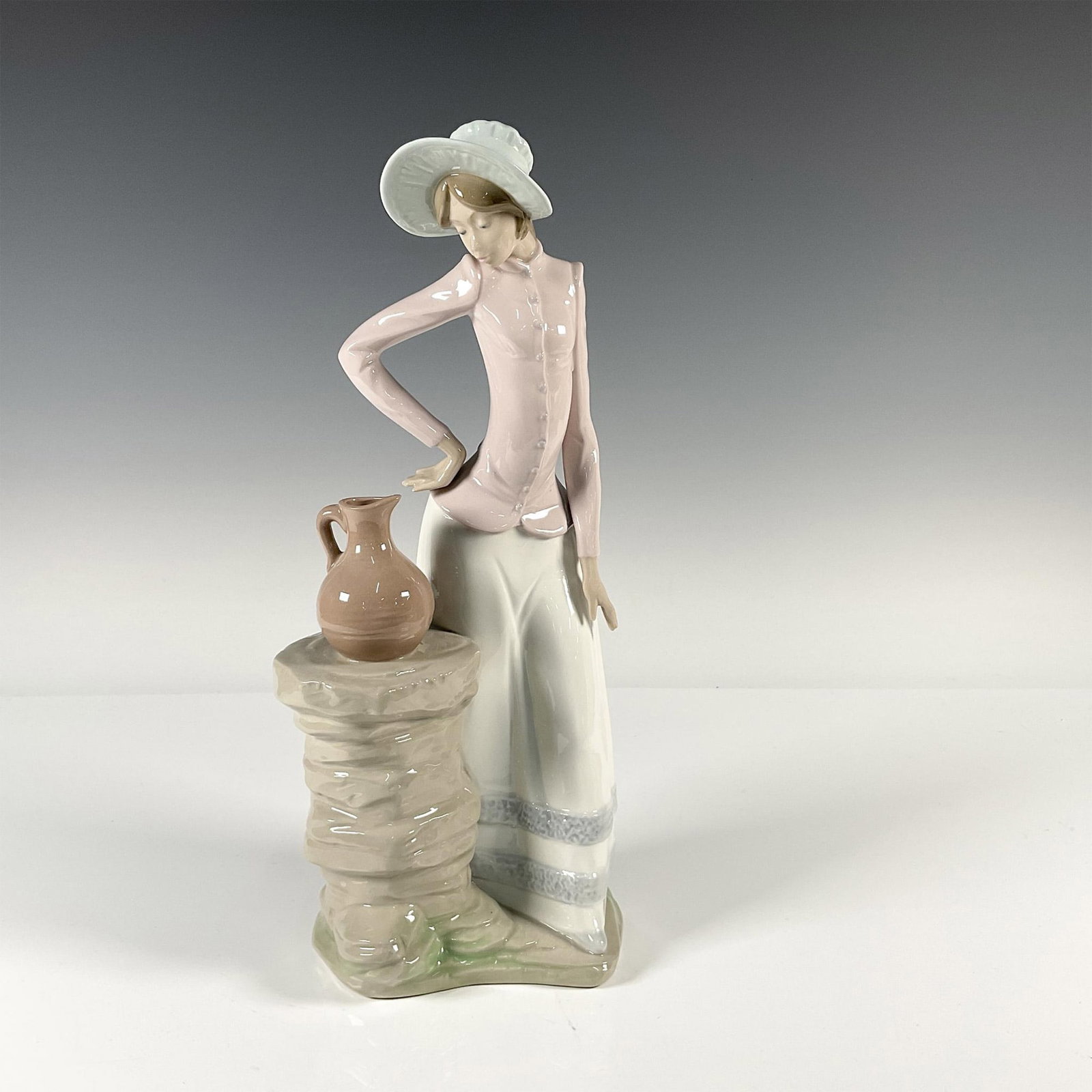 Nao by Lladro Porcelain Figurine, Woman With Pitcher: Woman eyeing a pitcher sitting on a stone pedestal. Gloss finish. NAO backstamp. Issued: 20th century Dimensions: 4"L x 5.25"W x 13"H Manufacturer: Nao by Lladro Country of Origin: Spain