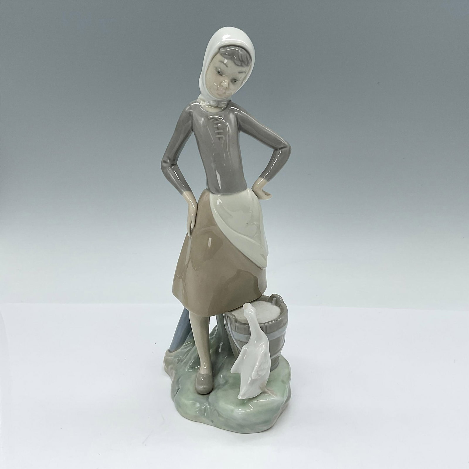 Girl with Milk Pail 1004682 - Lladro Porcelain Figurine: Glossy porcelain figurine depicts a girl standing with hands on hips, wearing a headscarf, with a duck and a bucket of milk at her feet. Lladro backstamp. Artist: Vincente Martinez Issued: 1970-1990 D