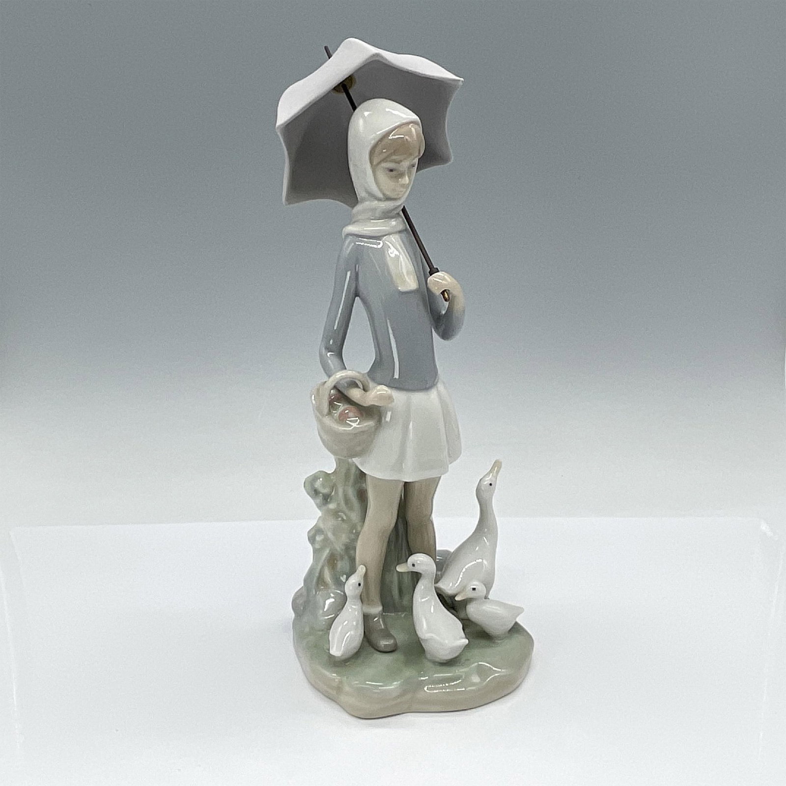 Girl with Umbrella and Geese 1004510 - Lladro Porcelain Figurine: A tall, glossy figurine featuring a girl with a basket in hand and an umbrella, gently observing a family of geese gathered around her feet. Lladro backstamp. Artist: Fulgencio Garcia Issued: 1969-199