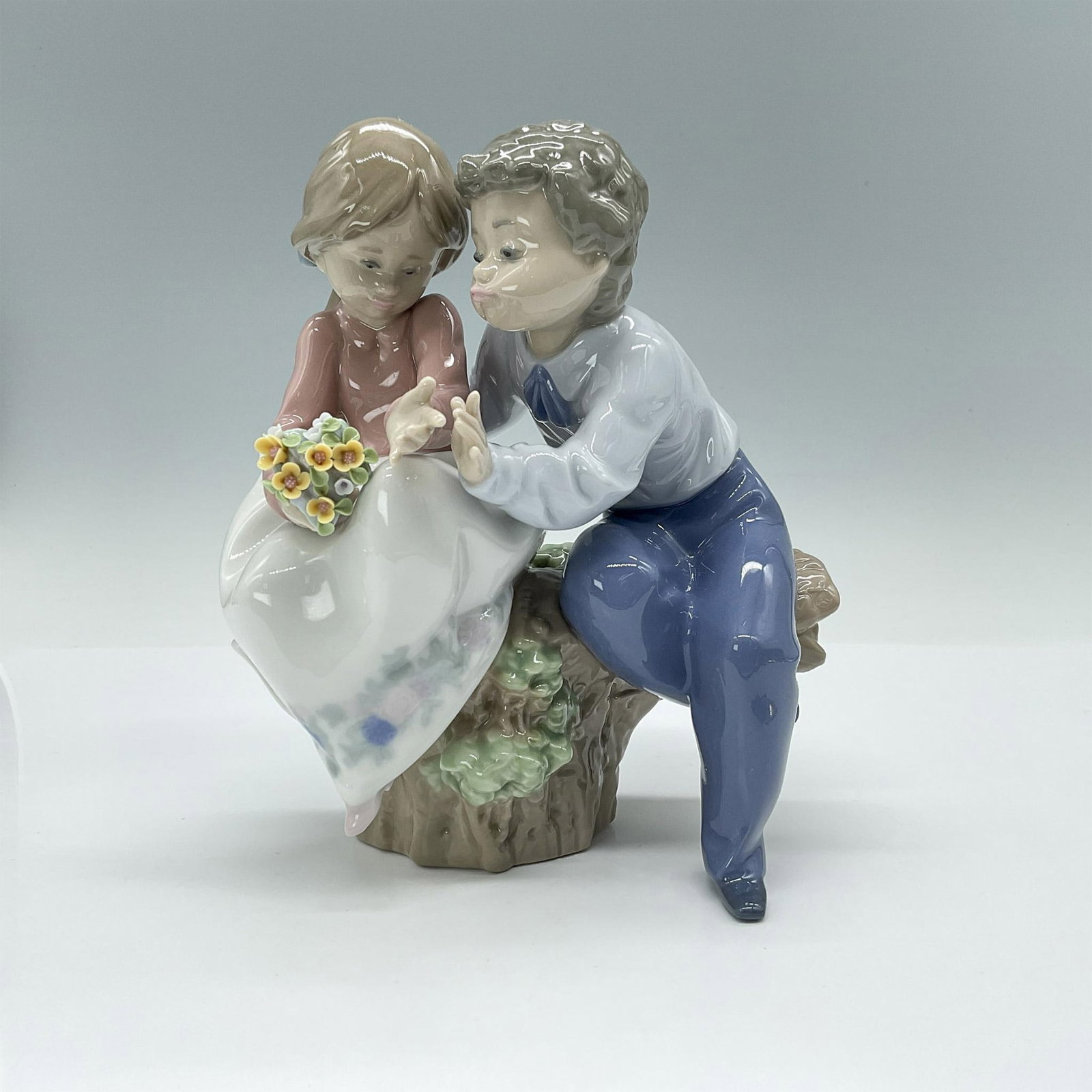 Just a Little Kiss 1005701 - Lladro Porcelain Figurine: Charming glossy figurine depicting two young people sitting on a tree stump. Lladro backstamp. Artist: Antonio Ramos Issued: 1990-1997 Dimensions: 6"L x 4.5"W x 7"H Manufacturer: Lladro Country of Or