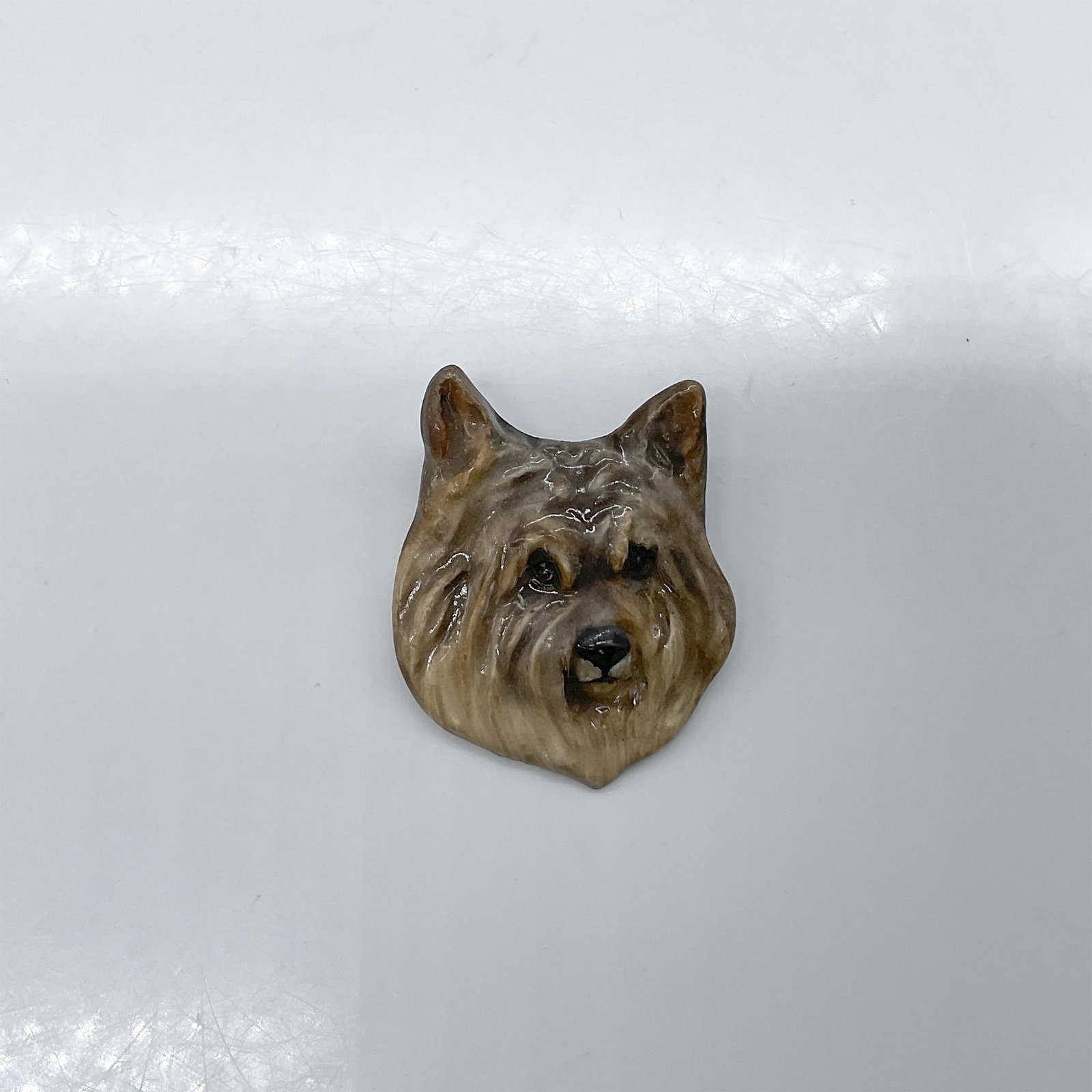 Royal Doulton Dogs Head Brooch, Cairn Terrier: Pottery pin molded as a terrier head; needle pin fasten. Gray and pale brown coloration. Royal Doulton impressed stamp. Issued: 1930s Dimensions: 1.25"W x 1.75"H x 0.5"D Manufacturer: Royal Doulton Co