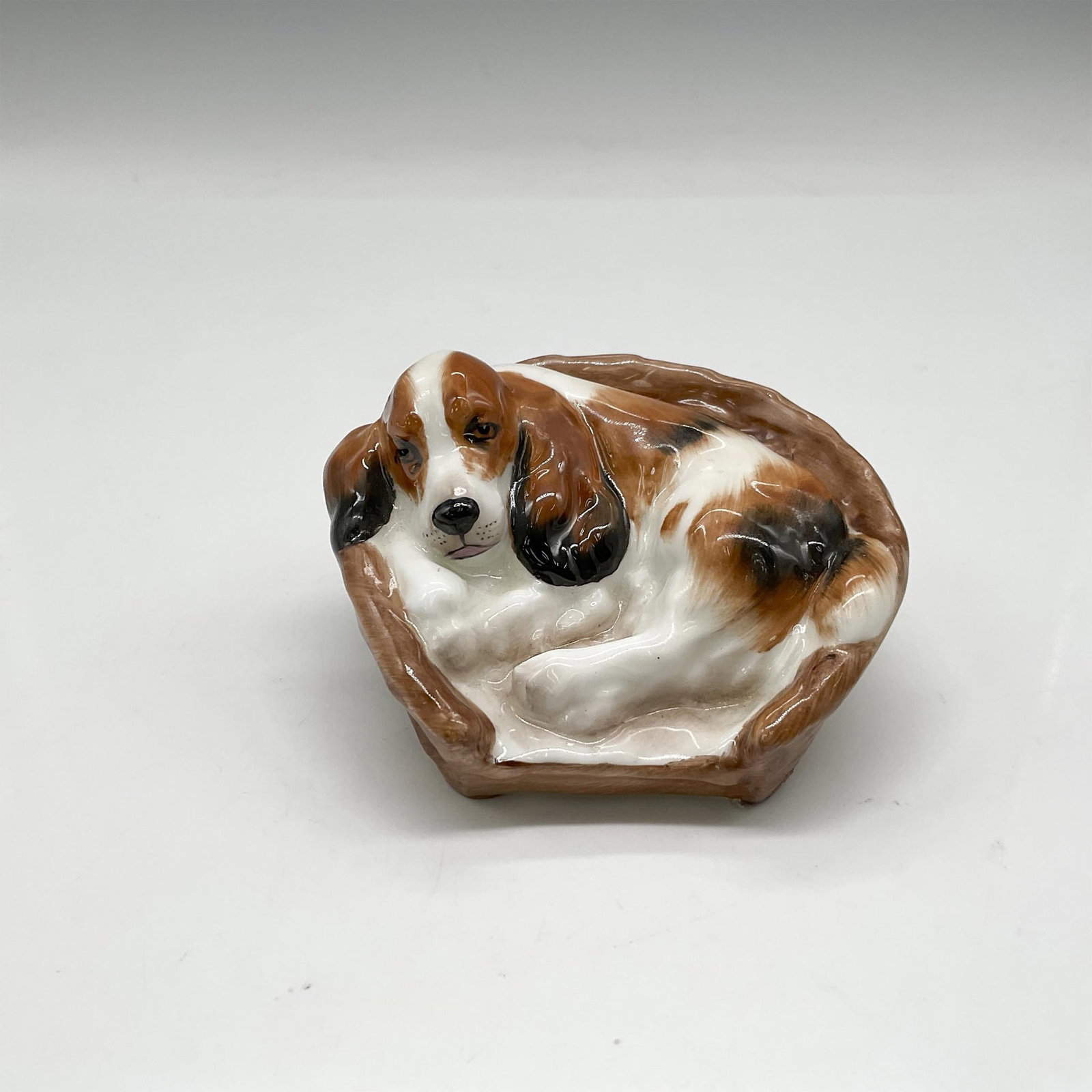 Royal Doulton Bone China Figurine, Cocker Spaniel HN2585: Dog relaxing in comfy basket is hand painted in white, brown and black. Royal Doulton backstamp. Issued: 1945 - 1985 Dimensions: 2.25"H x 3.5"W Manufacturer: Royal Doulton Country of Origin: England