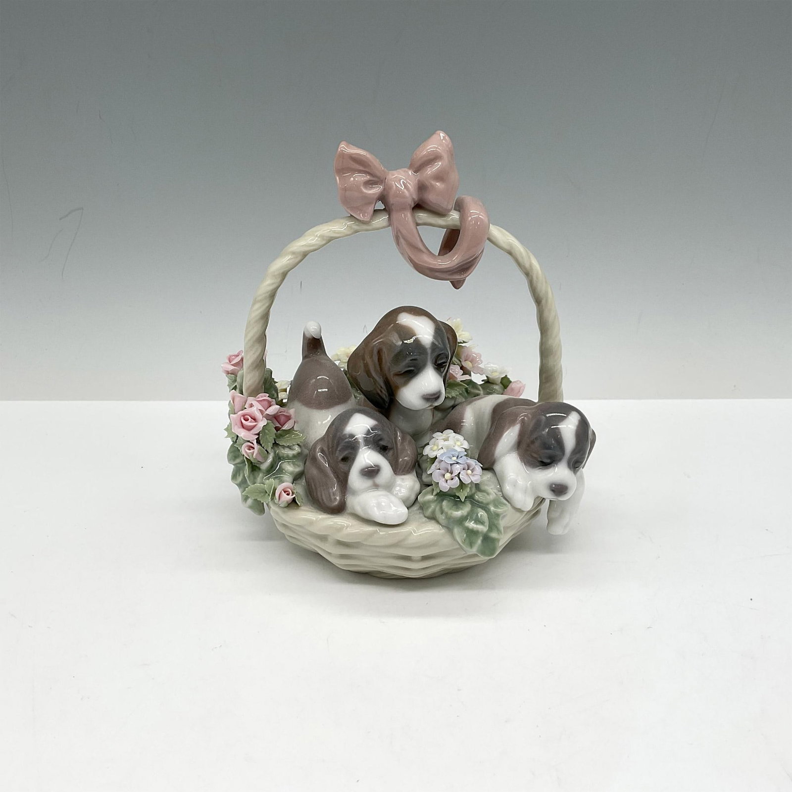 Litter of Love 1001441 - Lladro Porcelain Figurine: Designed to pull on your heart strings, this basket of puppies does it. Woven basket with rose colored ribbon at top holds 3 puppies that have brown patches over a white coat with colorful flowers aro