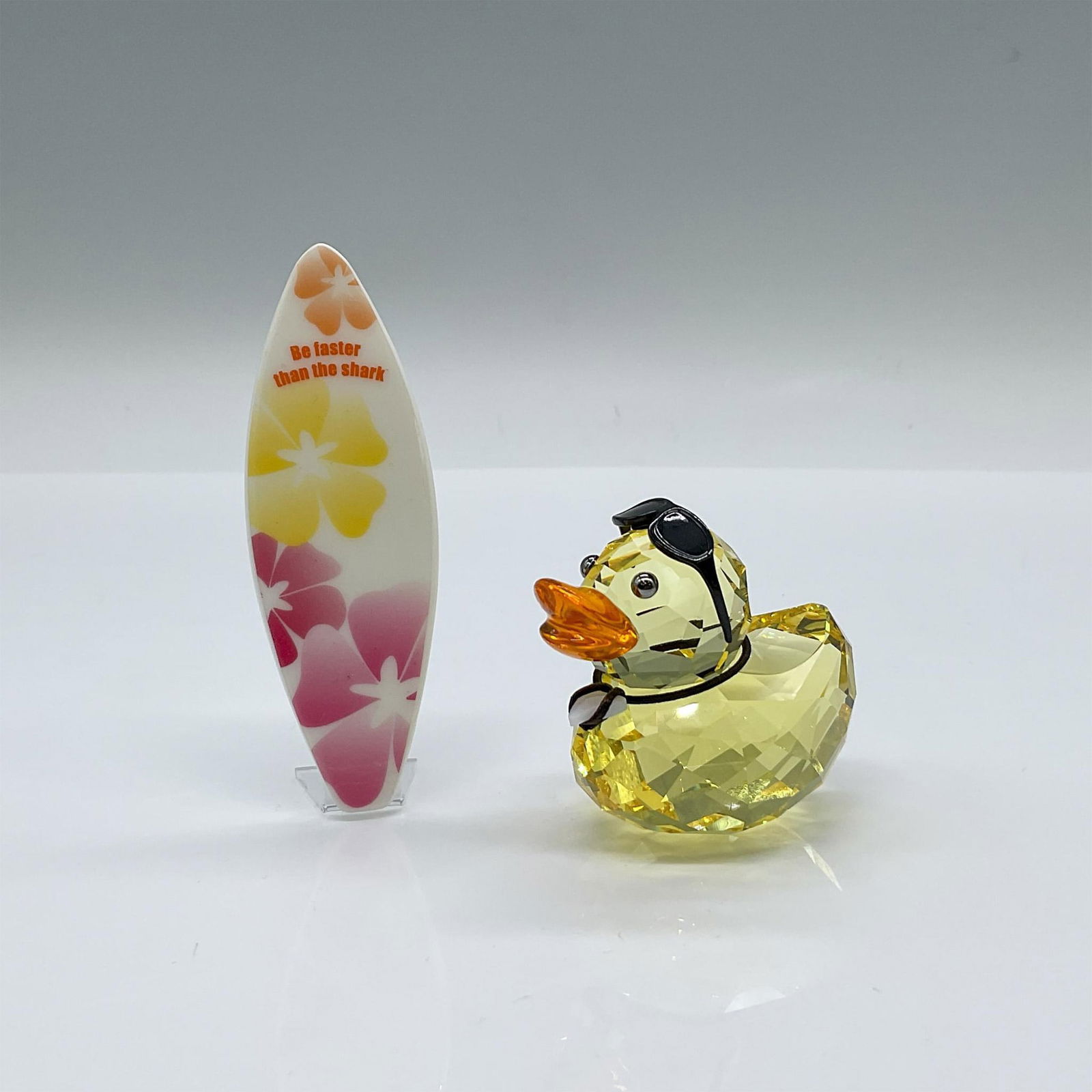 Swarovski Crystal Figurine, Happy Duck - Sunny Steve: A light topaz crystal duck with a sun crystal beak and silver metallic eyes. Sunny Steve comes with black metal removable sunglasses and a floral surfboard. He is wearing a white alabaster crystal sha