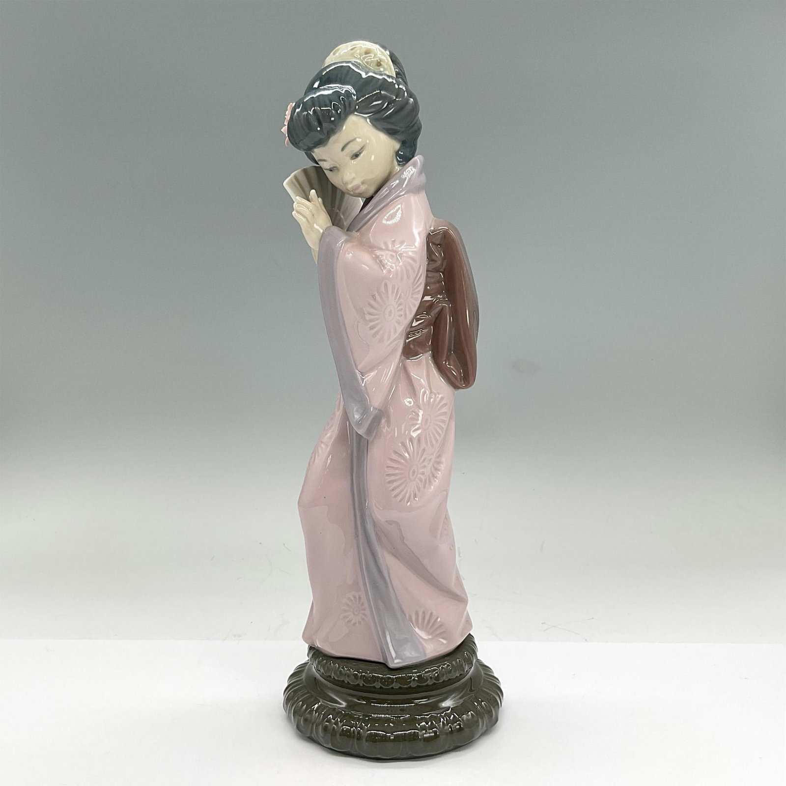Lladro Porcelain Figurine, Chrysanthemum 1004990: Glazed porcelain figure of a girl in a pink kimono holding a fan with flowers in her hair. Lladro backstamp. Dimensions: 4"L x 4"W x 11.5"H Manufacturer: Lladro Country of Origin: Spain