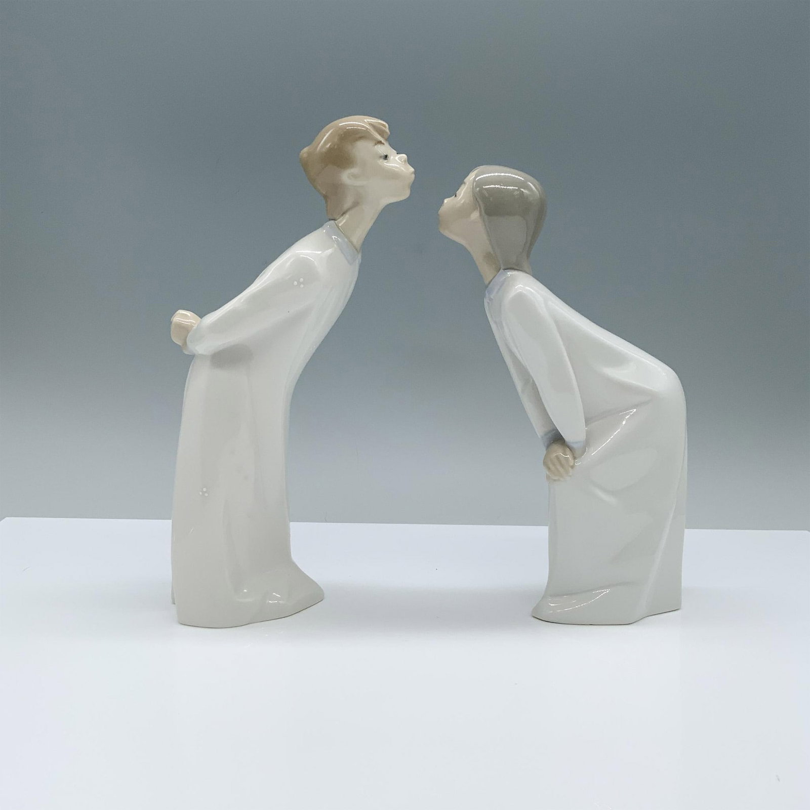 Lladro Porcelain Figurine, Kissing Girl and Boy: Lladro Porcelain Figurine, Kissing Girl 1004873 and Boy 1004869. A glazed porcelain figurine featuring a girl and a boy, both adorned in flowing white robes, delicately bent forward with puckered lips