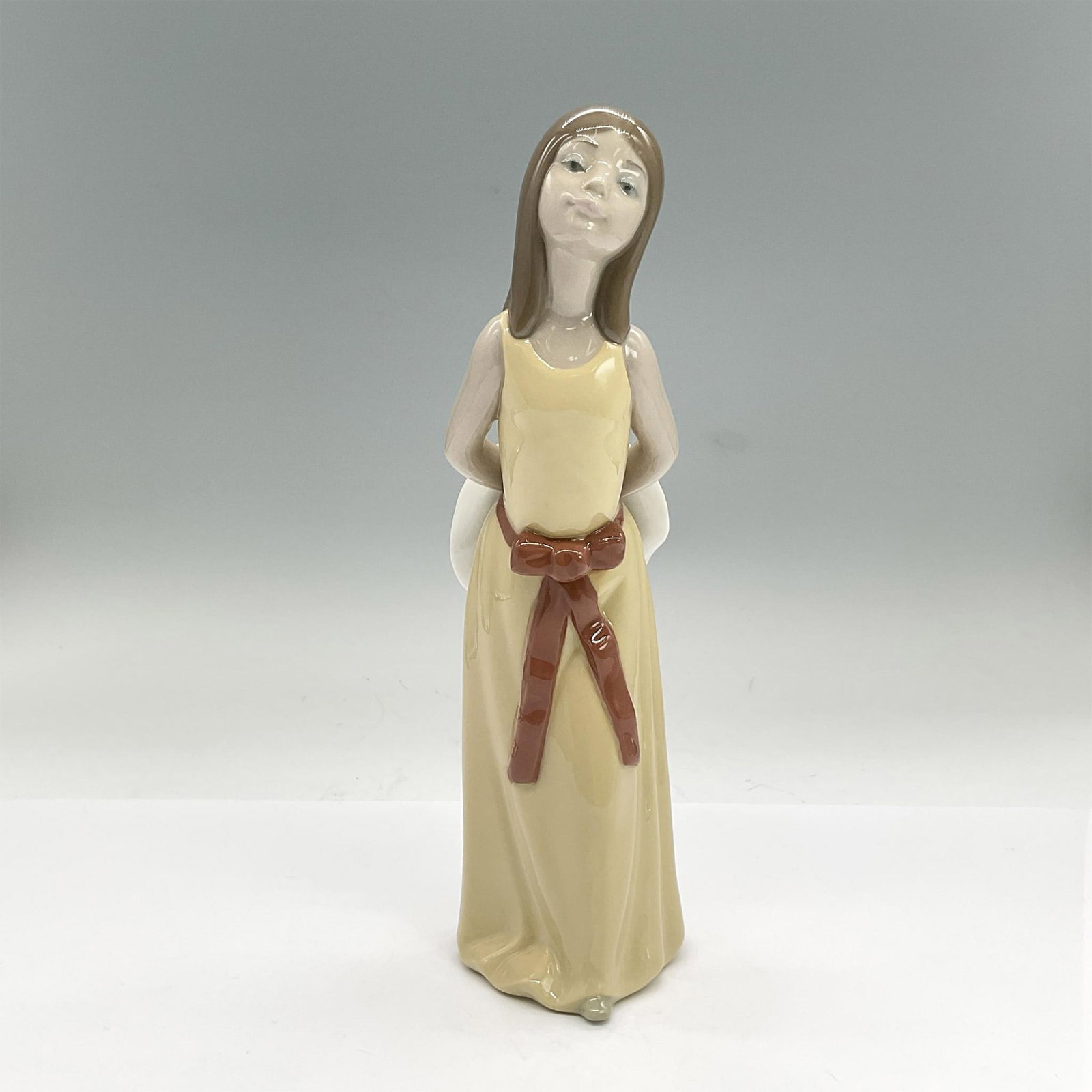 Lladro Porcelain Figurine, Naughty 1005006: Glazed porcelain figure of a girl in a yellow dress holding a wide brimmed hat behind her. Lladro backstamp. Issued: 1978-1998 Dimensions: 3"L x 2.5"W x 9.5"H Manufacturer: Lladro Country of Origin: S